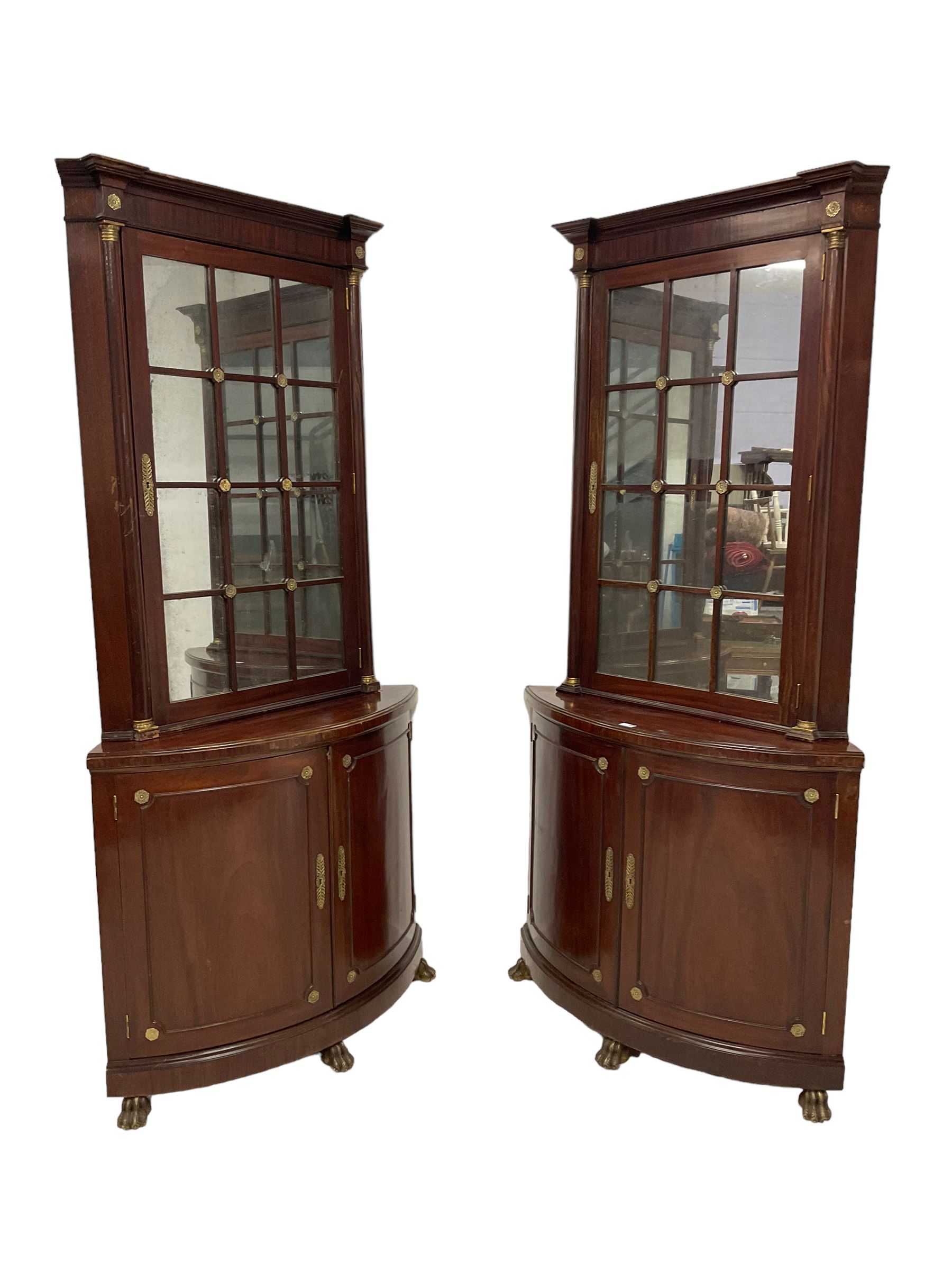 Pair of French Empire design corner cabinets, the straight-front upper section with canted corners and applied half-column pilasters, enclosed by mirror glazed door, the bow-front lower section enclosed by two doors with applied mouldings, mounted by cast metal floral rosettes, skirted base with cast gilt metal hairy paw brass feet