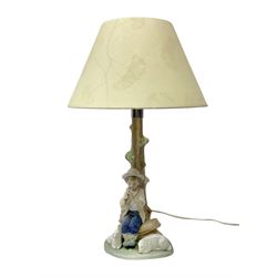 Nao table lamp modelled as a boy playing the flute upon logs with two recumbent sheep at his feet, the stem modelled as a tree trunk, with fabric shade, with printed mark beneath, H46cm
