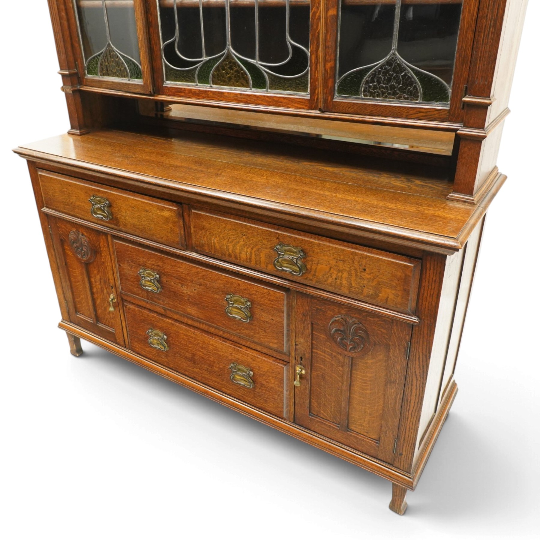 Arts and Crafts oak dresser, projecting moulded cornice above three glazed panels with coloured leaded glass floral panels, flanked by two glazed doors enclosing two adjustable shelves, open mirrored recess below with mirrored back, the base fitted with two short drawers over two central drawers flanked by carved panelled cupboard doors, ornate brass handles, raised on tapering square legs