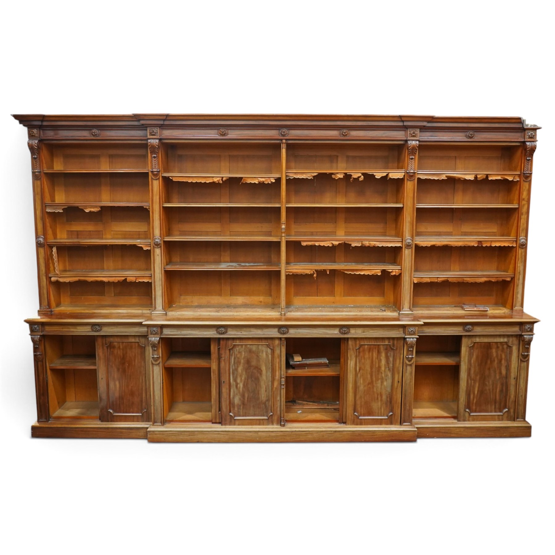 Large early 19th century Country House mahogany breakfront library bookcase, the projecting moulded cornice over a frieze applied with carved flowerhead paterae, the open panelled upper section fitted with adjustable shelves and shaped valances, divided by moulded pilasters with acanthus-carved corbels, the projecting base enclosed by eight panelled cupboard doors with central turned canted pilaster, on plinth base