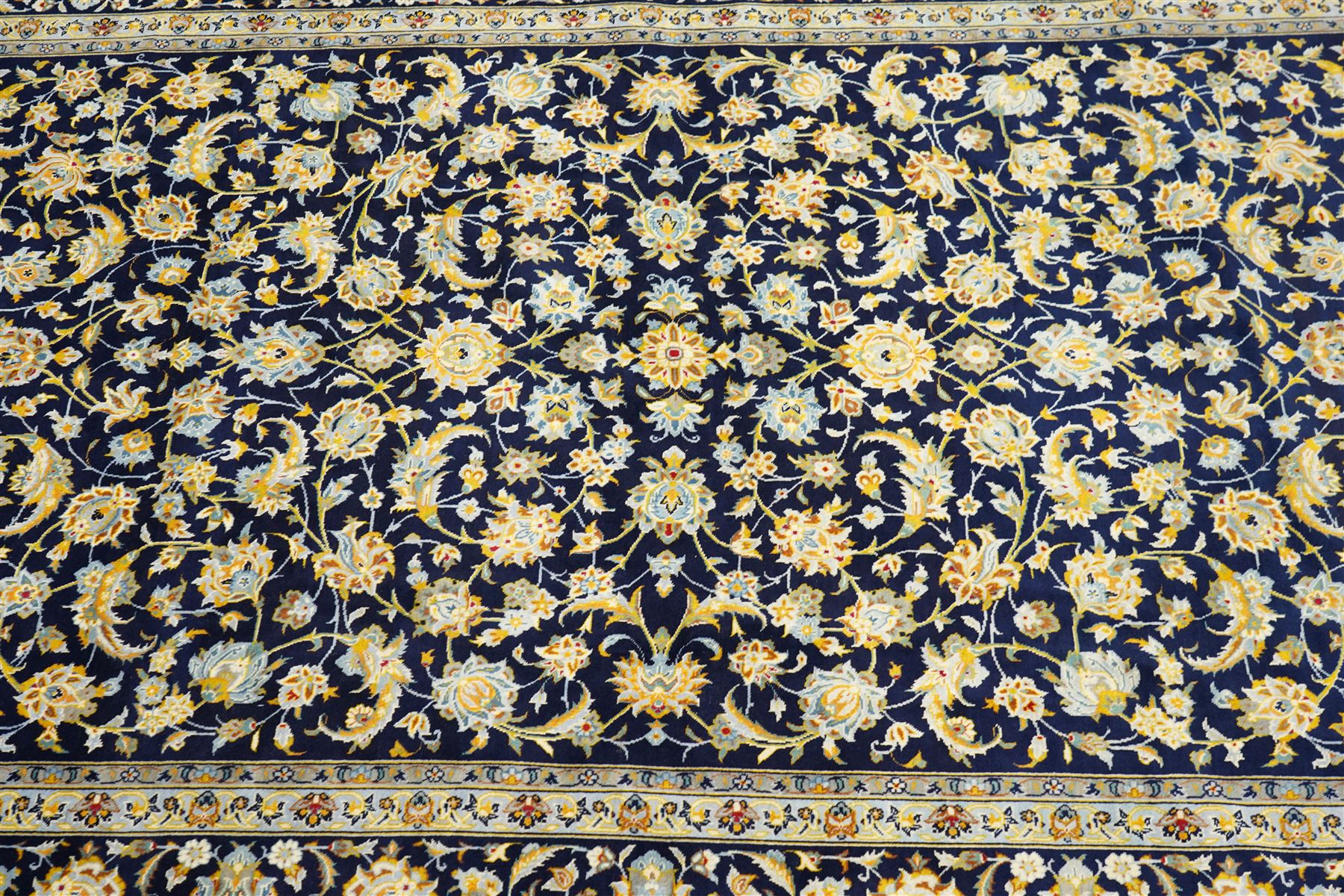Central Persian Kashan hand-knotted carpet, deep indigo field with a densely all-over arrangement of scrolling floral vines, palmettes, and flowering sprays, in soft ivory, pale blue, and warm gold tones, principal ivory border with a continuous meandering vine of stylized palmettes, and floral motifs, flanked by multiple narrow guard borders, with cotton fringe ends 