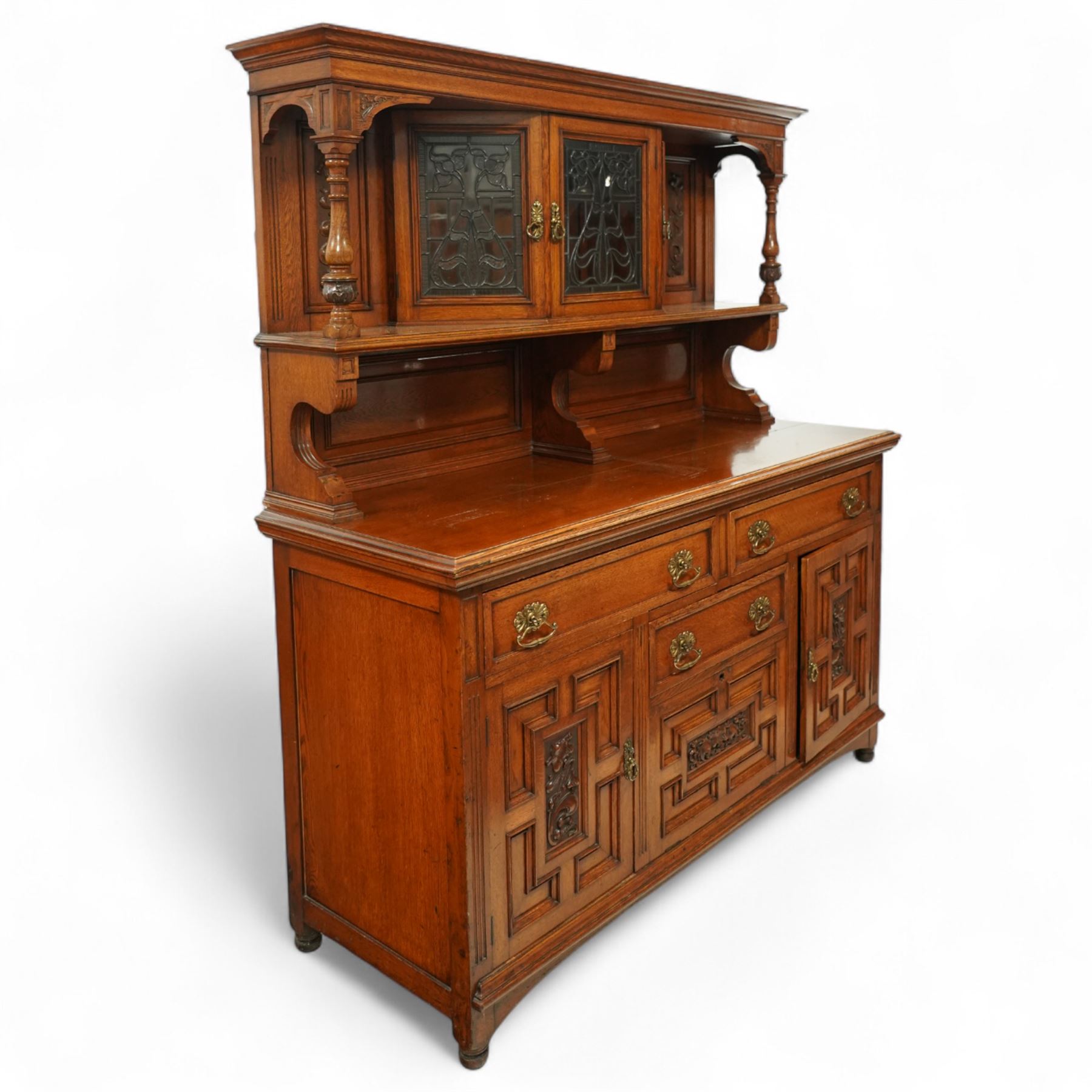 Early 20th century golden oak dresser sideboard, superstructure with three stained and leaded glass doors flanked by carved uprights above an open display shelf, the base with three drawers and geometric panelled cupboard doors, enclosing drawers with cutlery and bottle compartments, raised on short feet