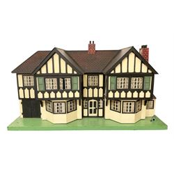 Large Tri-ang No.93 doll`s house in the 'Stockbroker's Range' c1935; two-storey double-fronted Tudor design, half timbered gables, two-storey bay windows, cream and mock shrubbery to front, opening metal framed windows with green shutters, red simulated tiled roof with chimney, integral garage with opening doors, front elevation has four hinged doors opening to reveal two bedrooms with fire places, living room, bathroom and kitchen with built in dresser and sink, entrance hall and staircase, side entrance with porch and seat and sun dial over on chimney stack; some wiring for electrical lighting; metal Tri-ang Toys disc verso; comprehensively furnished H66cm L117cm D43cm