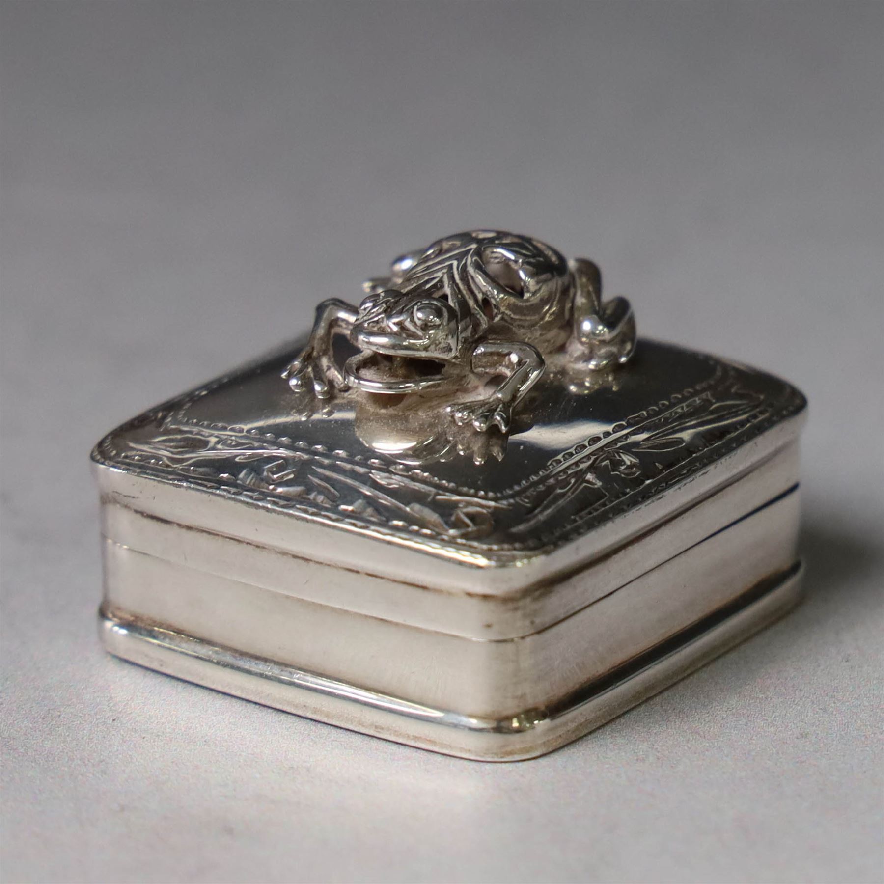 Modern silver pill box, of square form, the hinged cover surmounted with figure of a frog within a engraved scroll edge, hallmarked hallmarked Martin Gerald & Sid Adler, London 2000, W3cm