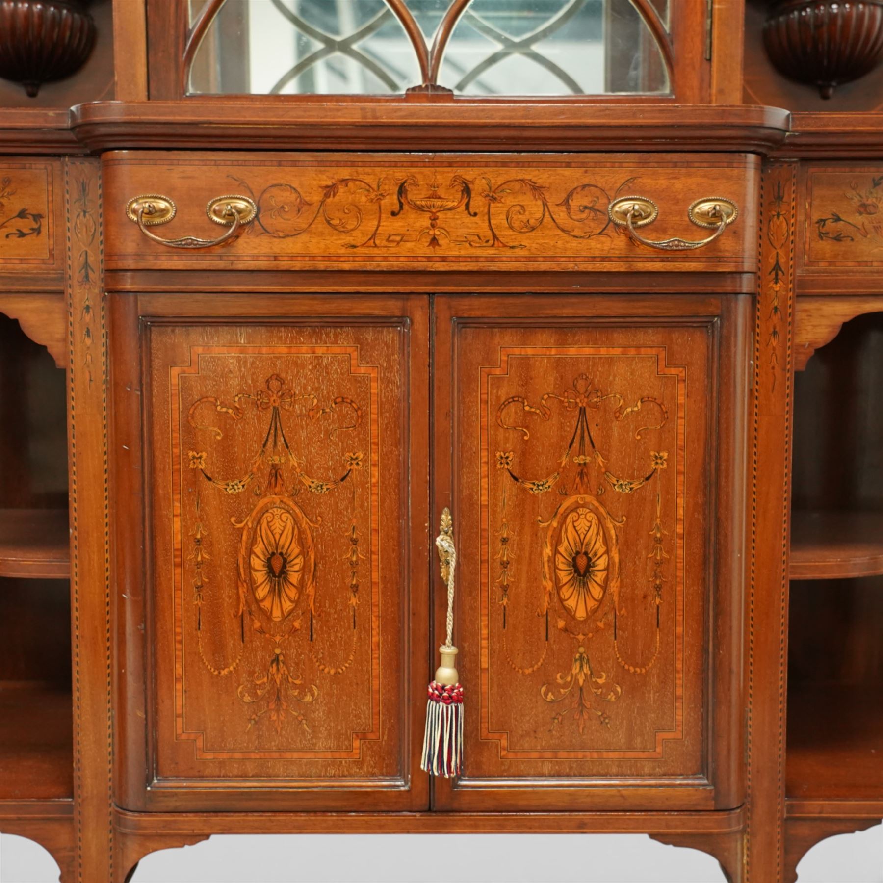 Edwardian Sheraton Revival inlaid mahogany breakfront cabinet, the broken swan neck pediment with central finial inlaid with cartouche and scrolling floral festoons, the central cabinet fitted with astragal-style glazed door with central urn and swag motif, flanked by arched bevelled mirrors over a shelf and gadrooned bracket over paterae inlays, base fitted with marquetry frieze rawer over a panelled cupboard with intricate inlaid scrolling floral designs, flanked by open shelves, raised on square tapering herringbone inlaid supports united by undertier