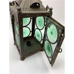 An Arts & Crafts wrought iron lantern, of square sided form with swing handle, and green and clear glass panels, raised upon four compressed bun feet, not including handle H30.5cm.