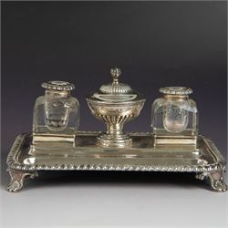 George IV silver desk stand, of rectangular form with rounded corners and oblique gadrooned rim, upon four embossed pad feet, with central pedestal dish of part fluted form with domed lid engraved with crest and with bud finial, with two recesses either side housing faceted glass inkwells, each with silver collars, hallmarked John & Thomas Settle, Sheffield 1825 & 1827, H11cm