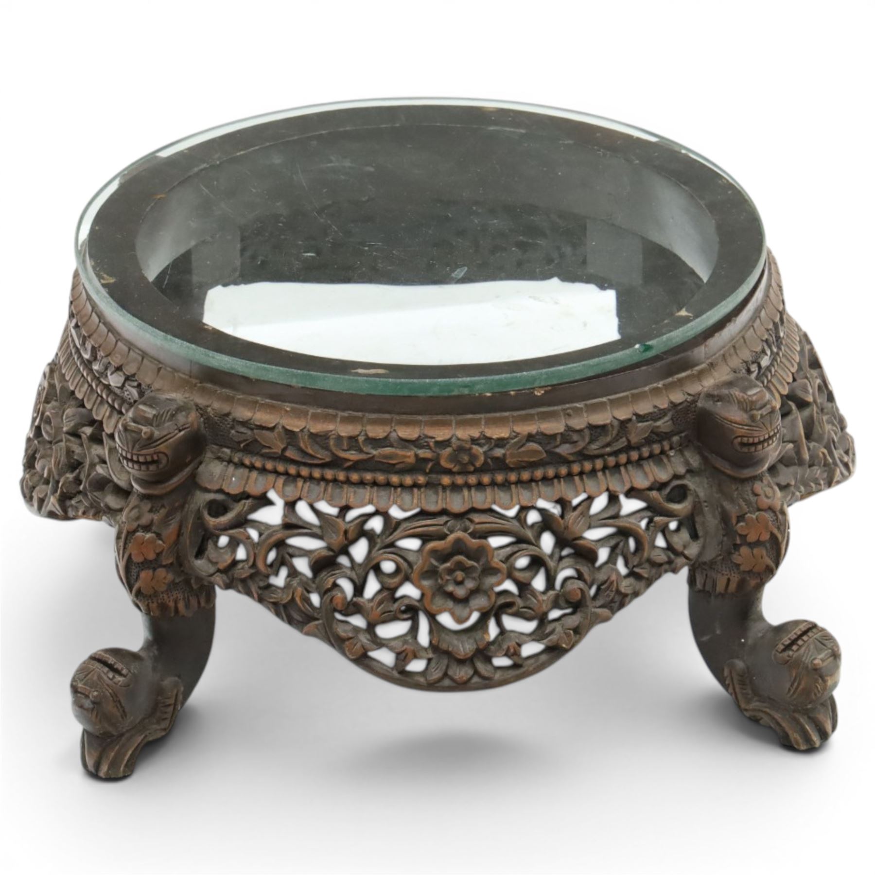 19th century Burmese carved hardwood jardinière stand, circular glazed top over pierced and carved floral frieze, on four zoomorphic supports carved with mythical beast masks and paw feet