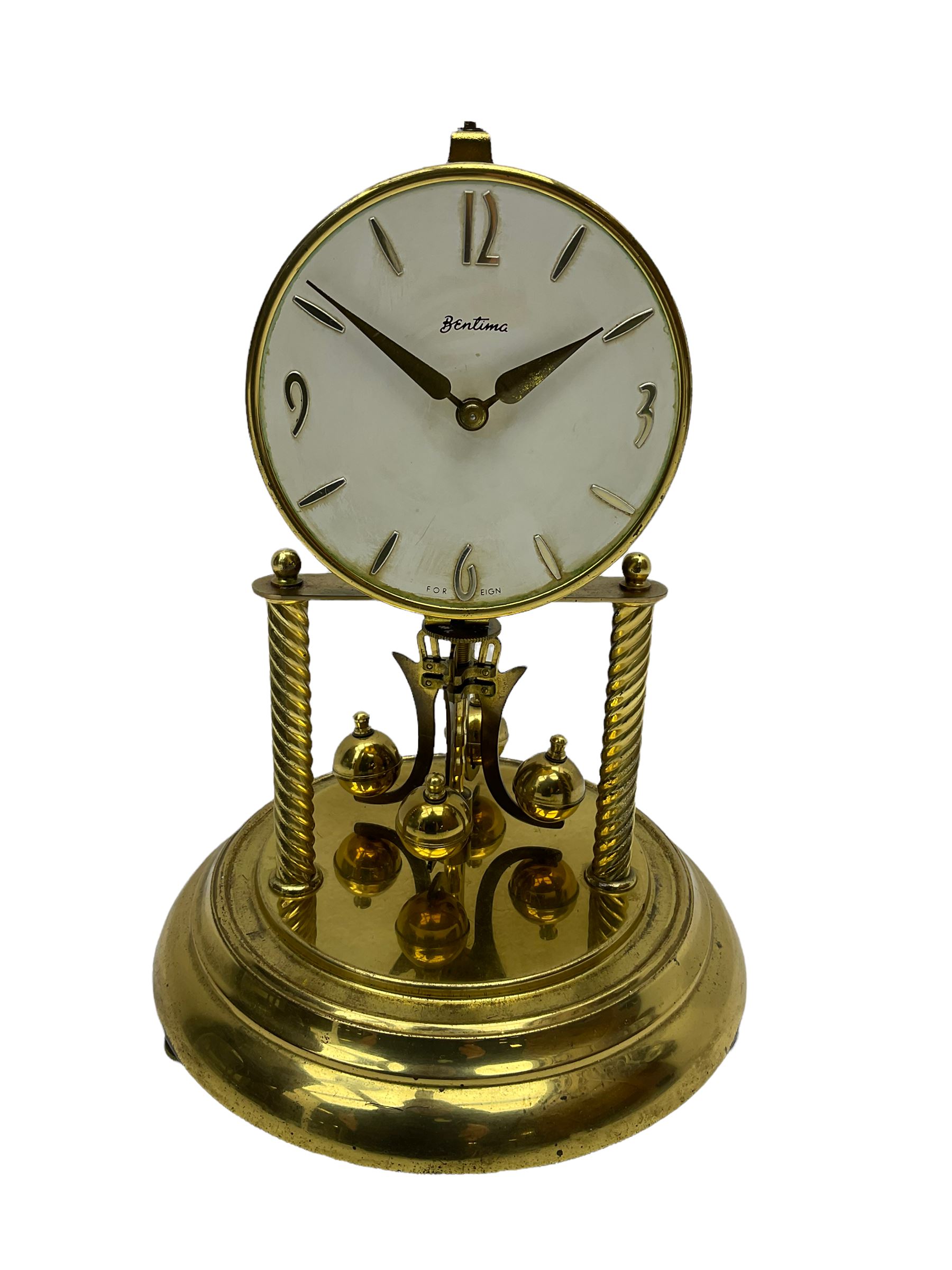 A mid-20th century “Bentima” 400-day anniversary torsion clock with a 7” circular brass base and unenclosed movement supported on two twisted columns, with a white dial gilt numerals and baton hands, four-ball rotary pendulum and glass dome. 

