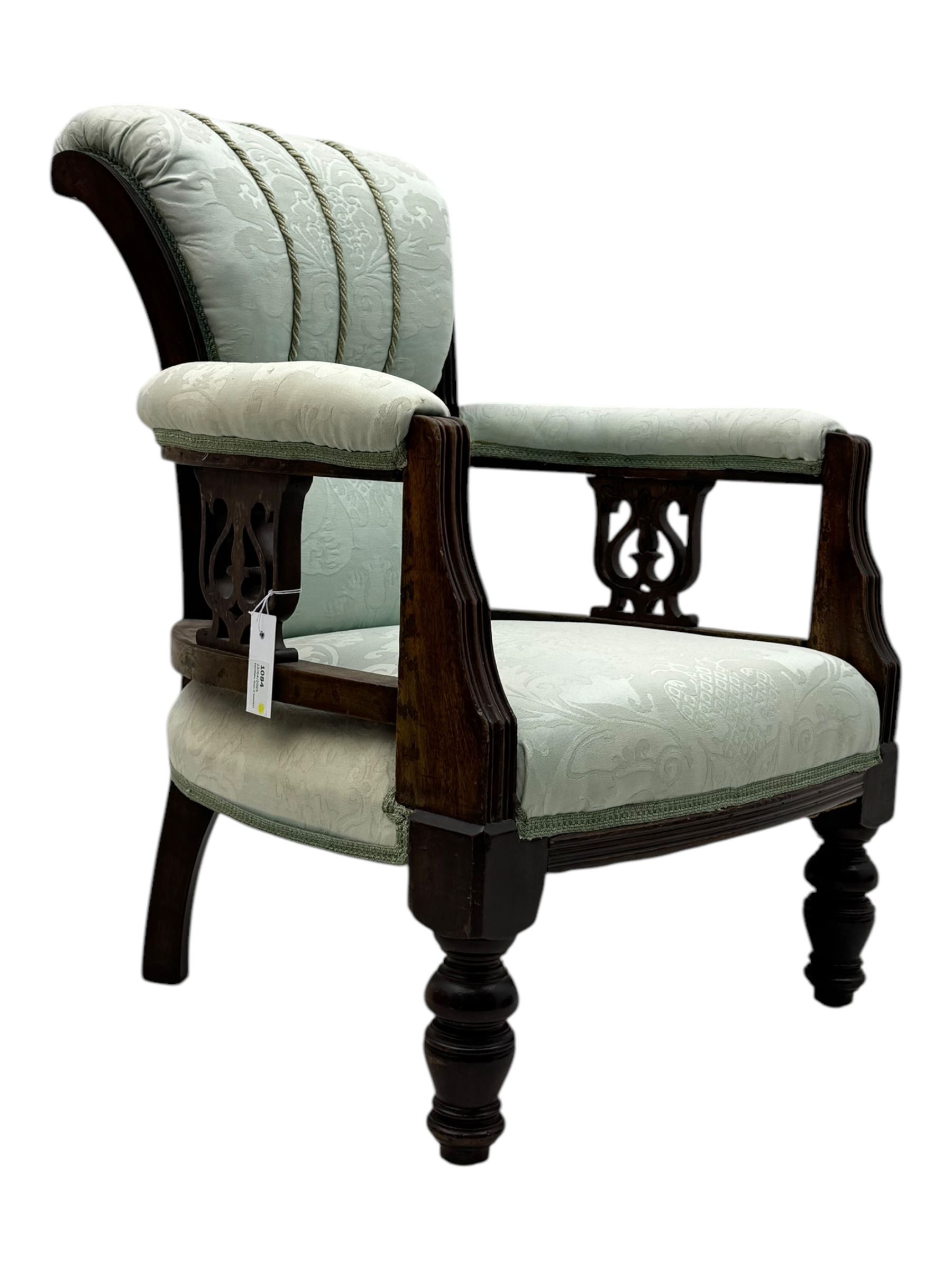 Late Victorian carved mahogany open armchair, the shaped back with fluted upholstered panel and padded arms above pierced scroll carved side supports, over a broad upholstered seat, on turned front supports