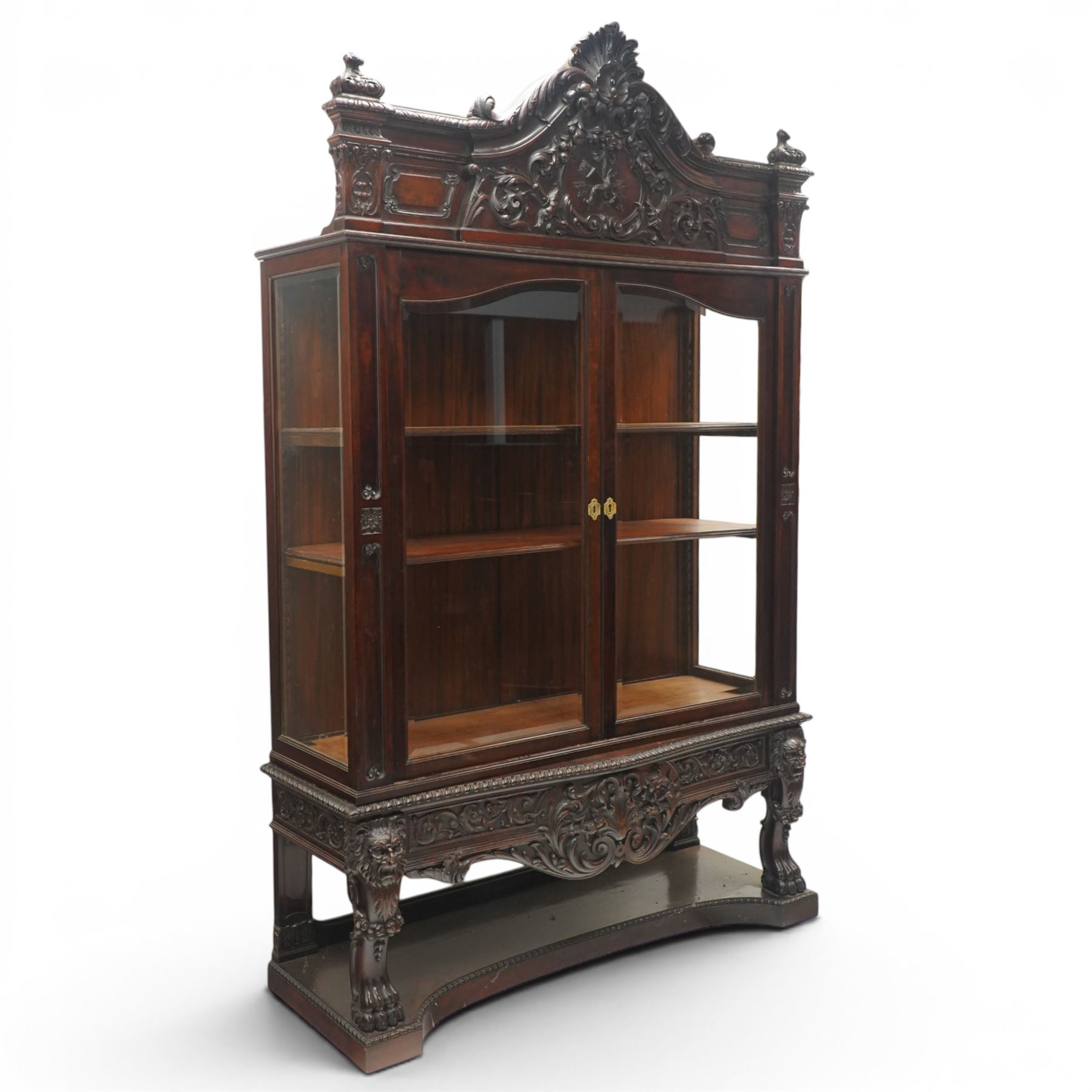 Large and impressive 19th century heavily carved mahogany display cabinet, the ornate pediment carved with a central scalloped and foliate rocaille motif with extending acanthus leaves and egg-and-dart moulding flanked by acanthi finials, over a central cartouche with carved bow-and-arrow motif and trailing scrollwork, the bookcase enclosed by two glazed doors with bevelled glass sides, the base with a gadrooned upper edge over a central rocaille ornament with scrolling acanthus leaves, the panelled frieze with further foliate motifs, raised on large carved snarling lion mask supports with floral garlands and claw feet, united by a concave rectangular platform base with gadrooning egg-and-dart moulding