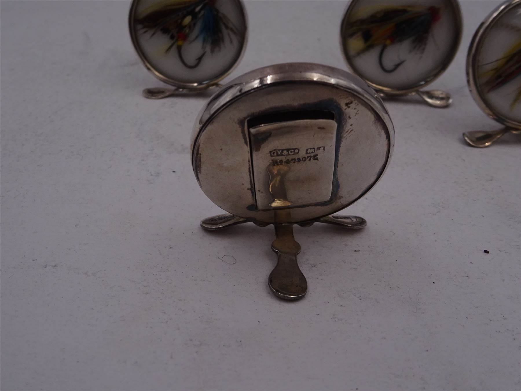 Set of four early 20th century silver mounted place card holders, each of circular form and set with a coloured fly fishing hook behind a glazed panel, upon three pad feet, hallmarked Grey & Co, Chester 1912, H4.5cm, contained within red fitted case, with purple silk and velvet interior