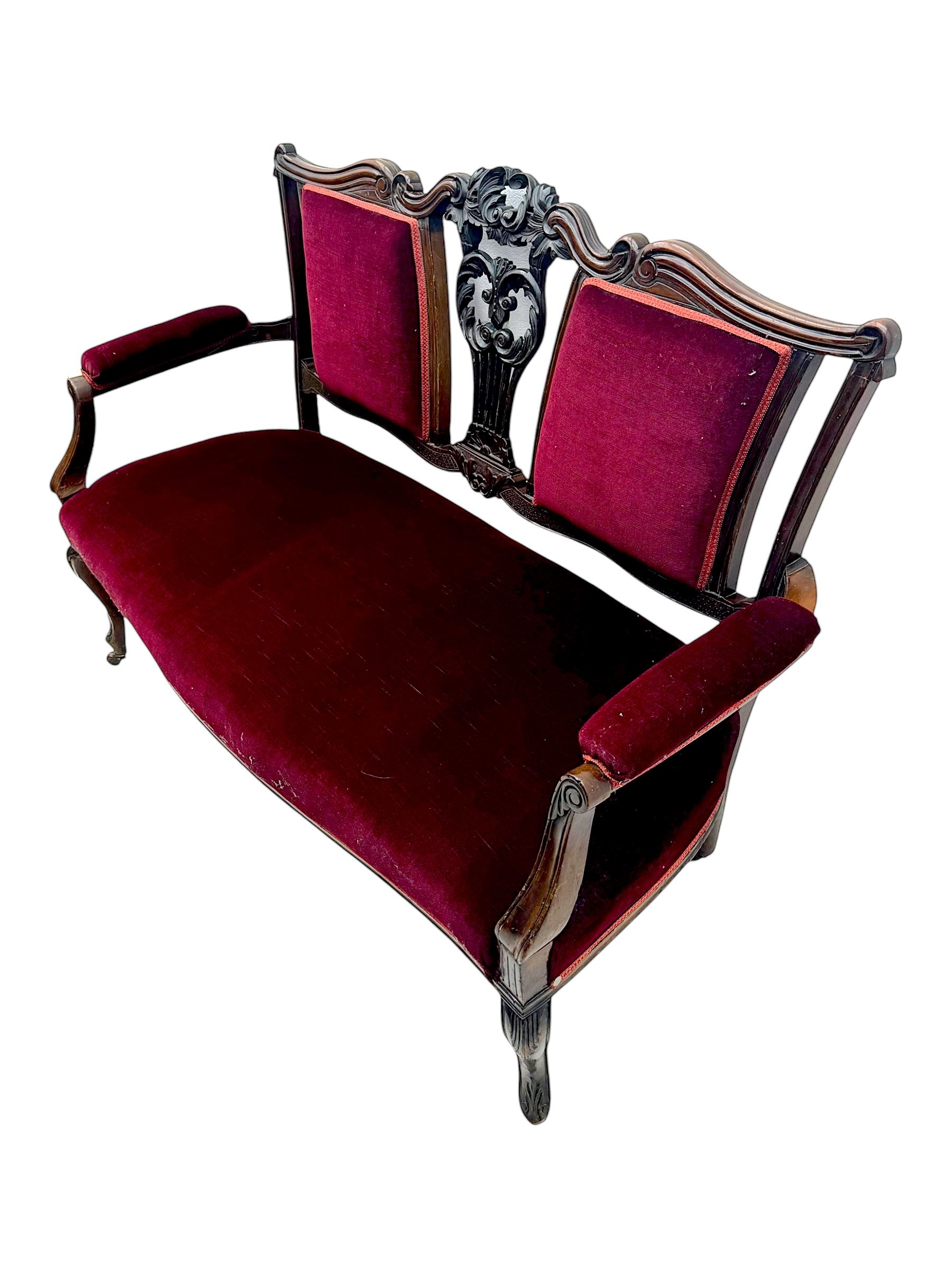 Late Victorian stained beech framed sofa, the backrests, seat and padded arms upholstered in deep red fabric, carved crest rail with scroll and foliate motifs supported by carved uprights, on cabriole supports with leaf carvings terminating in castors