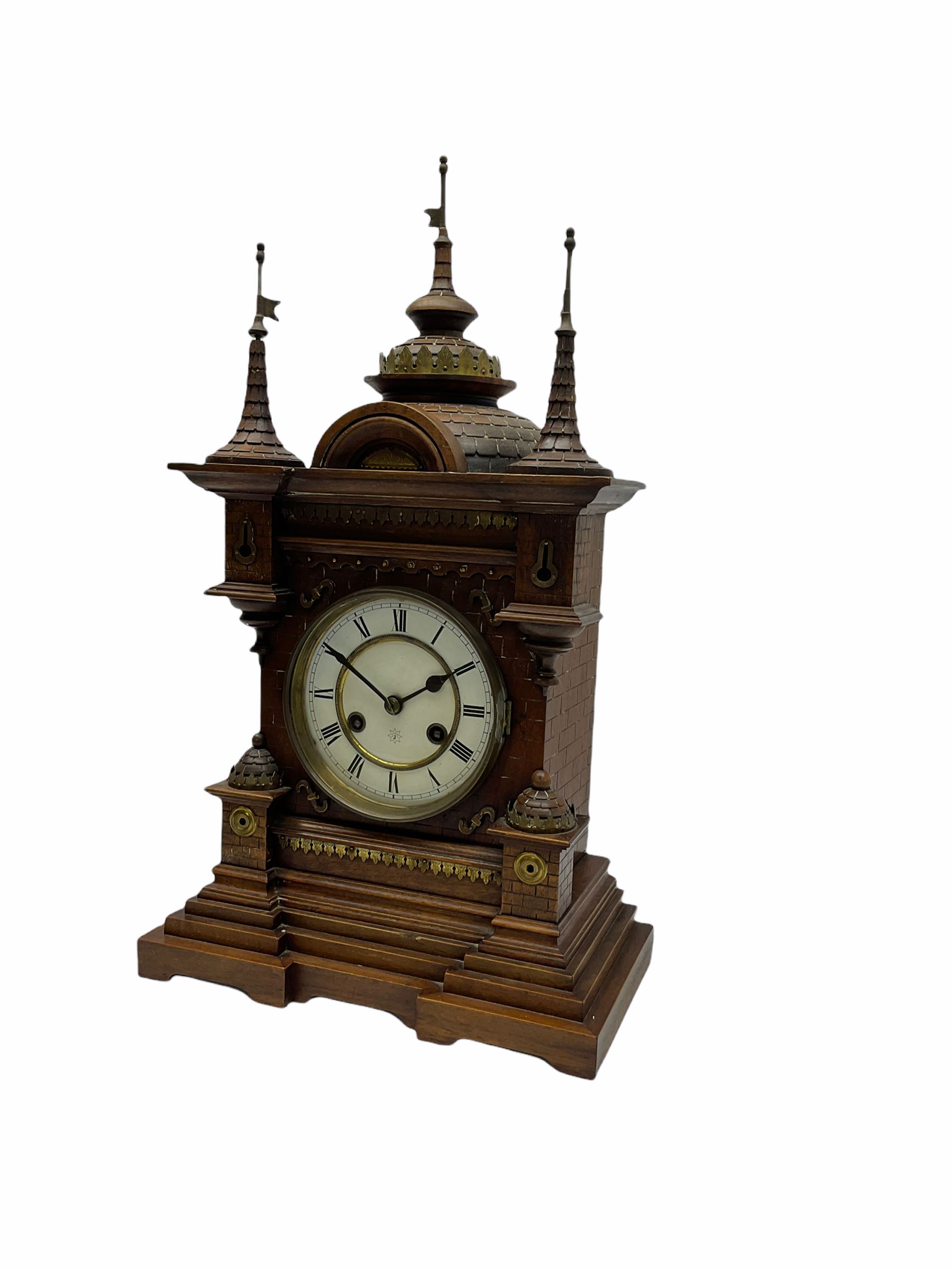 A late 19th century German Black Forrest mantle clock striking the hours and half hours on a coiled gong, two-piece white enamel dial inscribed with the six pointed Junghans logo, roman numerals and minute track, steel spade hands, spun brass bezel and glass, spring driven eight day movement with a recoil anchor escapement, carved oak case in fantasy form, multi-stepped plinth with incised brickwork effect to the sides and front of the case, with applied brass ornamentation, projecting turrets and gun ports, case surmounted with a pediment consisting of three distinctively carved oriental domes with roof tiles and guidon banner flags.
With pendulum.
