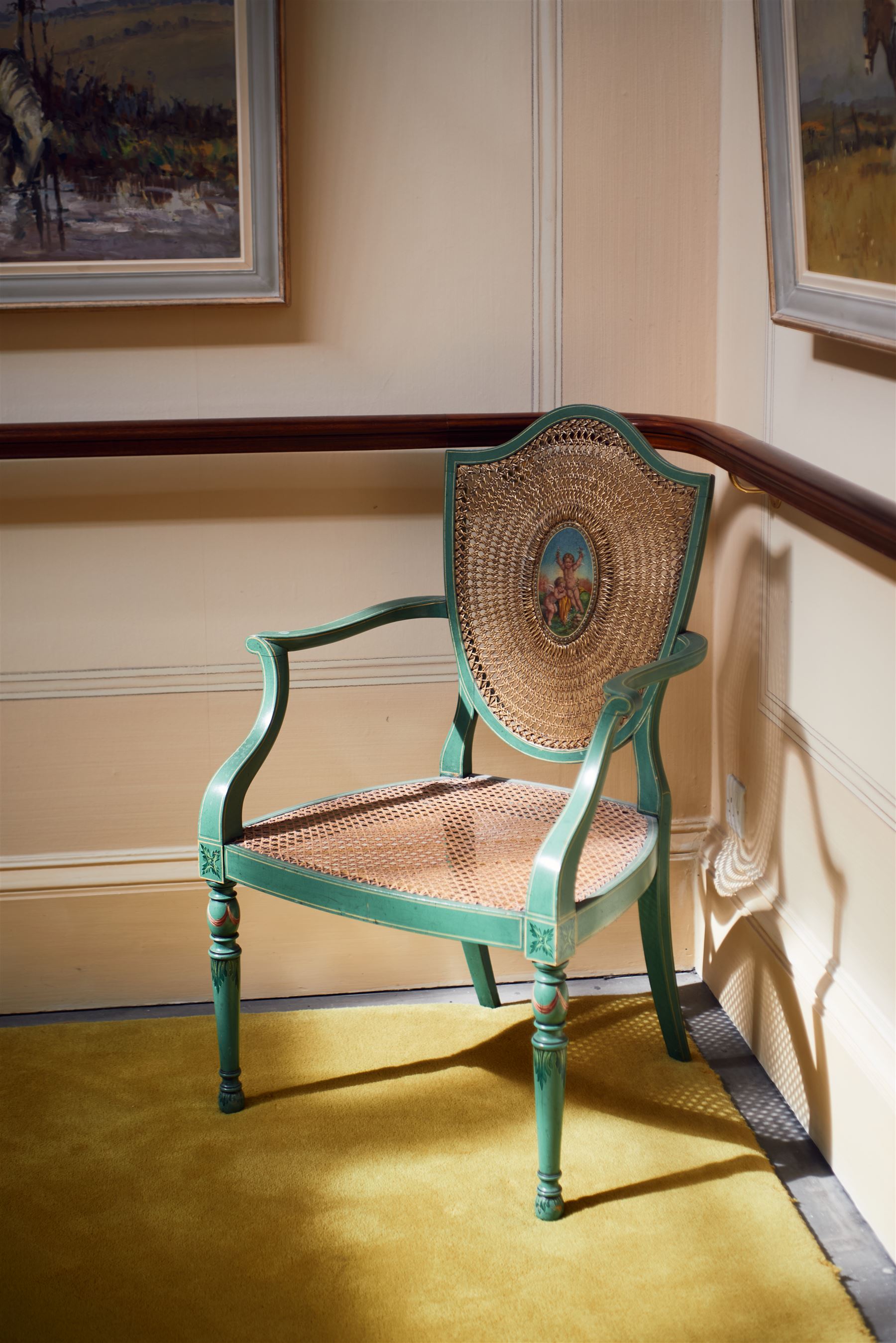 Pair of Edwardian Hepplewhite Revival green-painted open armchairs, cane work shield back with central oval panel panted with putto set upon rocky landscape with water and trees, shaped and rolled arms enclosing cane work seat, on turned front supports decorated with acanthus leaves and draped festoons 