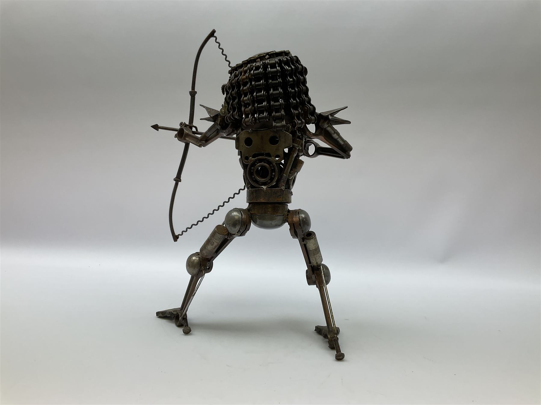 Predator metal sculpture, made with scrap metal, with articulated body, bike chain hair, and bow and arrow, H45cm