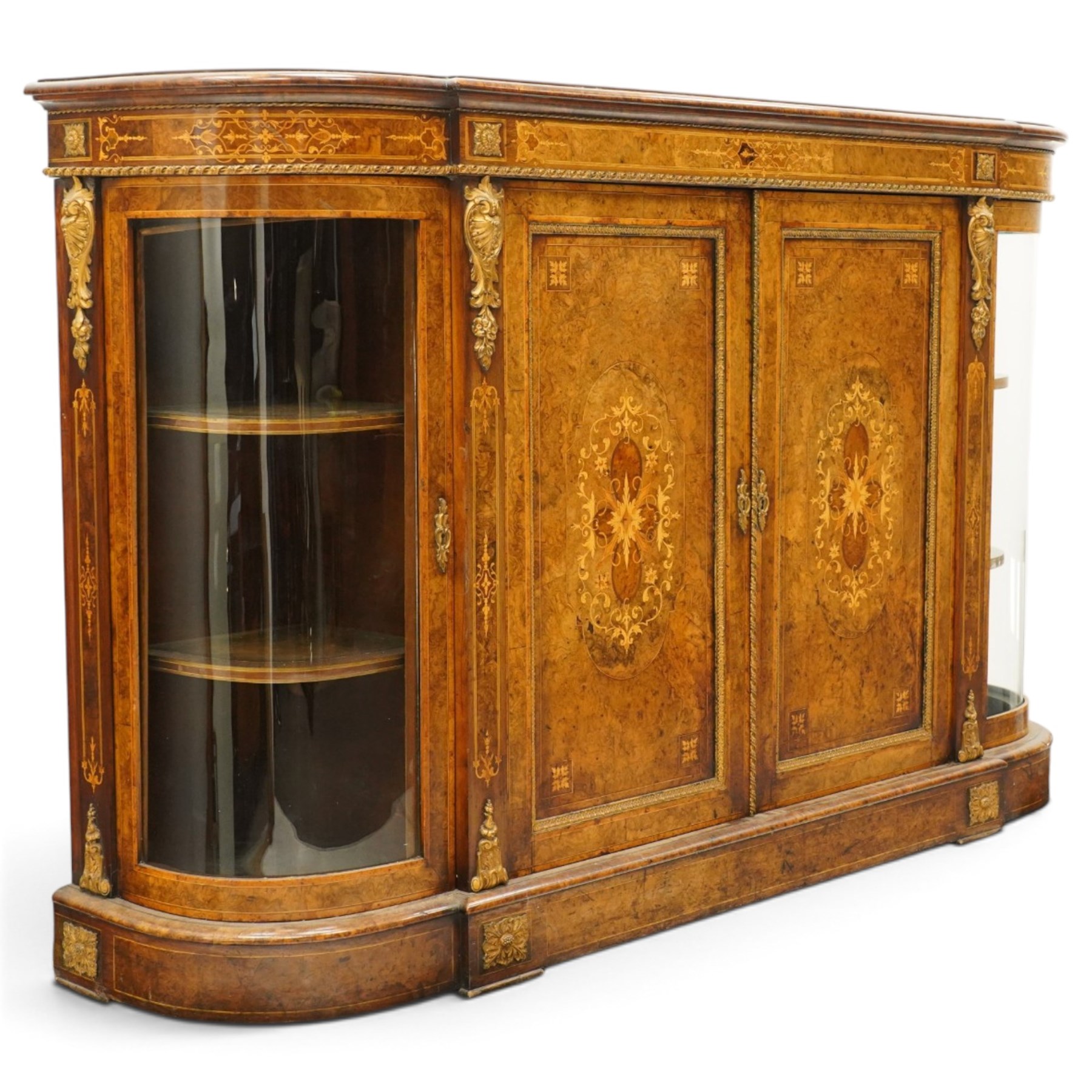 Large fine Victorian figured and burr walnut credenza, the breakfront D-shaped top with moulded edge, over an ornate frieze with inlaid Aesthetic Movement scrolling and geometric ornament, applied with gilt-metal ribbon-twist moulding and mounts, the central panelled doors profusely inlaid with scrolling foliate motifs and stringing, flanked by gilt-metal cartouche and scrolling acanthus corbel mounts, with flanking glazed bow-front side doors, on skirted base with further gilt metal foliate mounts