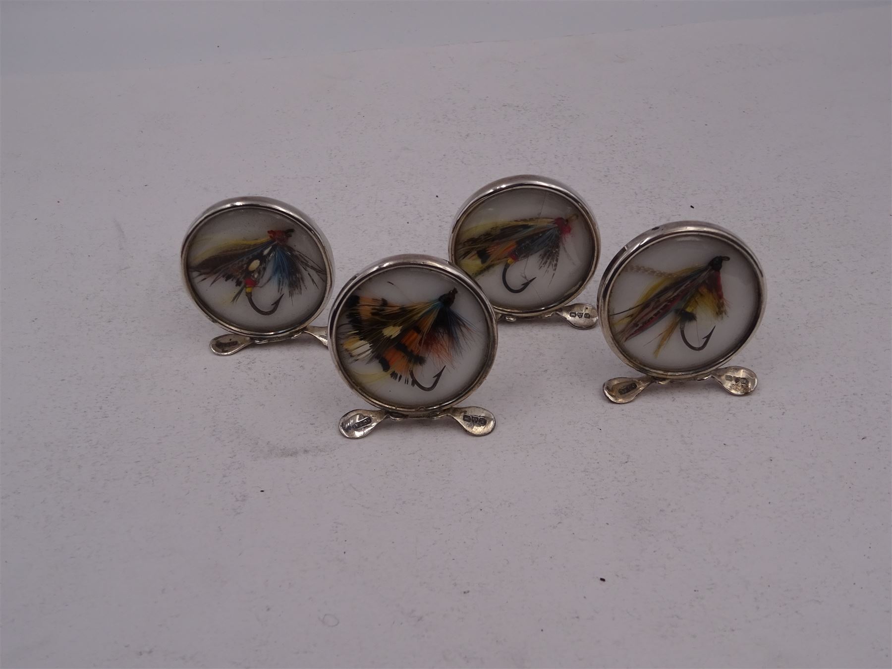 Set of four early 20th century silver mounted place card holders, each of circular form and set with a coloured fly fishing hook behind a glazed panel, upon three pad feet, hallmarked Grey & Co, Chester 1912, H4.5cm, contained within red fitted case, with purple silk and velvet interior