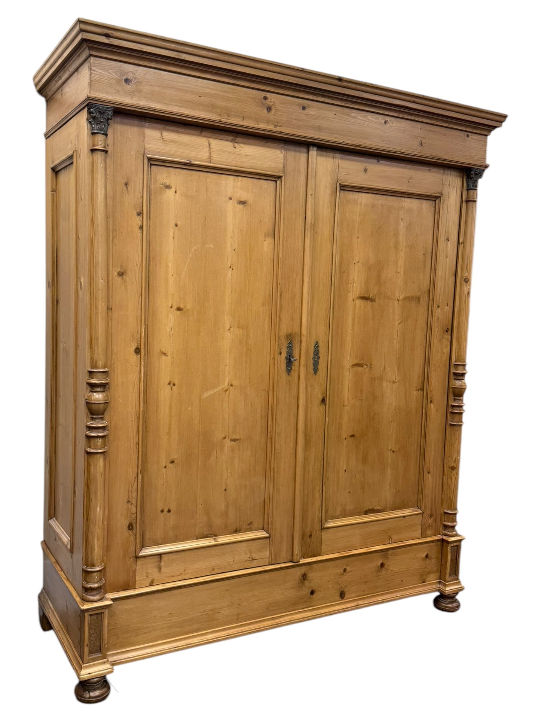 Large 19th-century Continental polished pine double wardrobe, panelled back and doors revealing brass hanging rod, flanked by half-turned columns with Corinthian capitals supported on bun feet