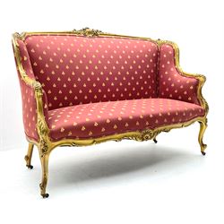 Victorian carved giltwood settee, the shaped cresting rail decorated with shell cartouche and flower head motifs, down swept arms with acanthus leaf and scrolled terminals, upholstered in red ground fabric decorated with repeating gilt motif pattern, cabriole supports with brass castors 