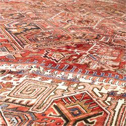 North West Persian Heriz hand-knotted carpet, madder red field, with a large angular stepped central medallion with serrated outlines, flanked by bold stylized palmettes, hooked leaves and geometric devices, spandrels similarly decorated within a wide geometric main border featuring angular floral motifs, enclosed by multiple guard borders in madder red, indigo blue, ivory, olive green and soft ochre, with cotton fringe ends 