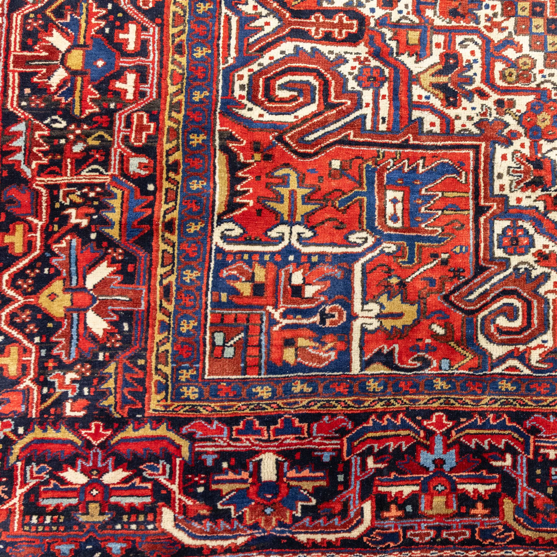 North West Persian Heriz hand-knotted carpet, madder red field, with a large angular stepped central medallion with serrated outlines, flanked by bold stylized palmettes, hooked leaves and geometric devices, spandrels similarly decorated within a wide geometric main border featuring angular floral motifs, enclosed by multiple guard borders in madder red, indigo blue, ivory, olive green and soft ochre, with cotton fringe ends 