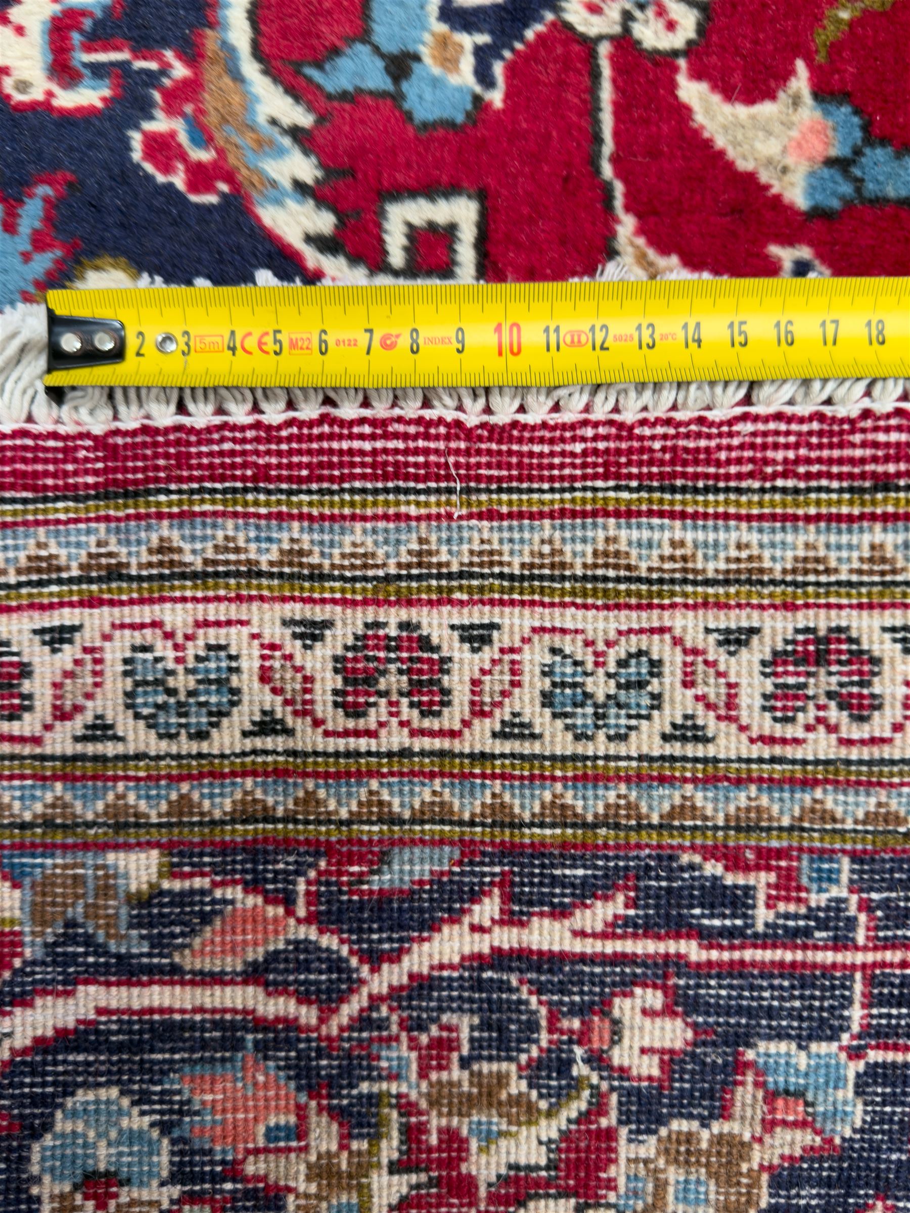 Central Persian Kashan hand-knotted carpet, the deep red ground with a central lozenge medallion in ivory and navy, surrounded by dense scrolling floral and vine motifs, navy spandrels and a wide border with stylised palmettes and flowerheads enclosed by multiple guard stripes