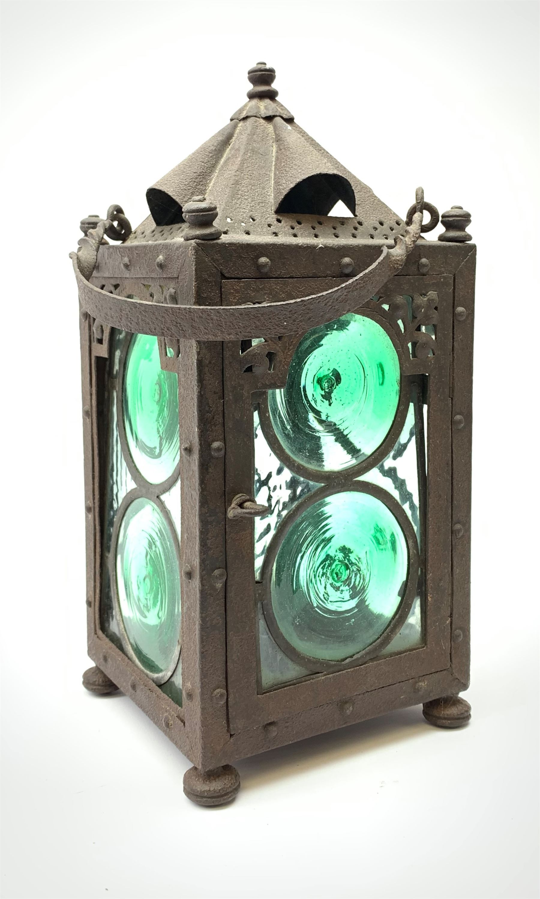 An Arts & Crafts wrought iron lantern, of square sided form with swing handle, and green and clear glass panels, raised upon four compressed bun feet, not including handle H30.5cm.