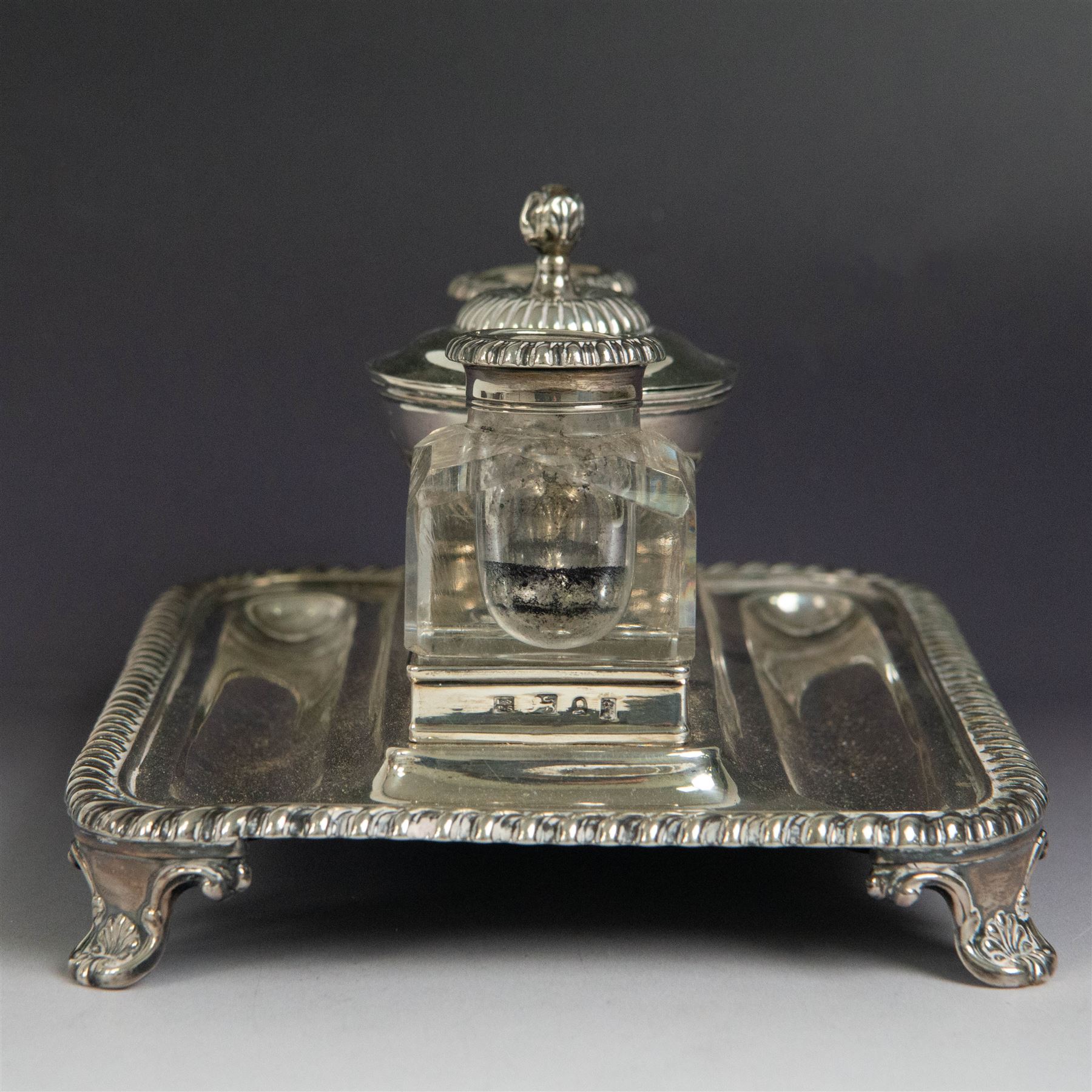 George IV silver desk stand, of rectangular form with rounded corners and oblique gadrooned rim, upon four embossed pad feet, with central pedestal dish of part fluted form with domed lid engraved with crest and with bud finial, with two recesses either side housing faceted glass inkwells, each with silver collars, hallmarked John & Thomas Settle, Sheffield 1825 & 1827, H11cm