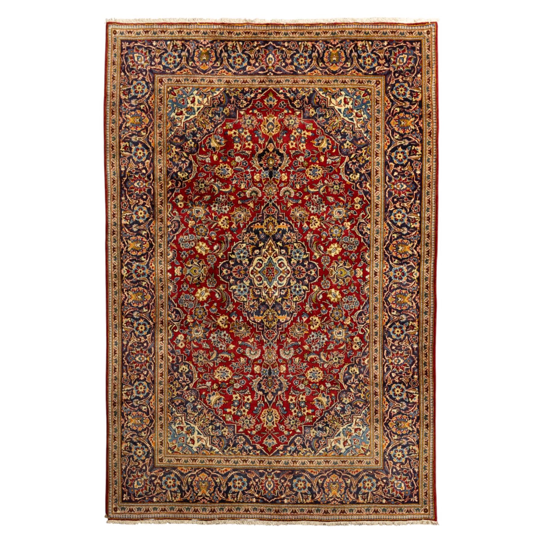 Central Persian Kashan hand-knotted carpet, crimson red central field with central floral medallion with floral vine-work and palmettes, within dense floral spandrels and a wide navy blue border decorated with continuous foliate motifs and multiple guard borders, with cotton fringe ends  