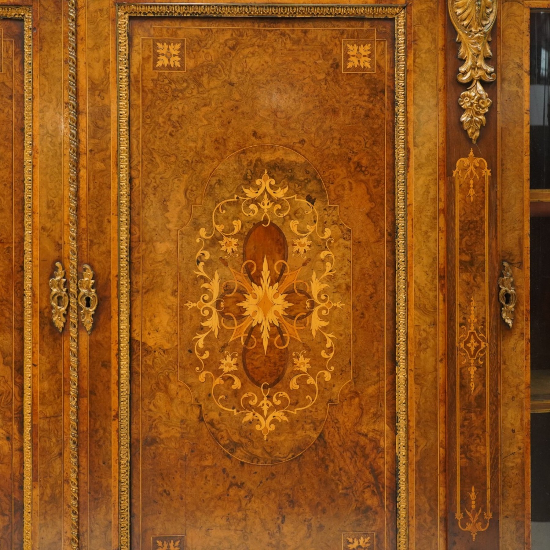 Large fine Victorian figured and burr walnut credenza, the breakfront D-shaped top with moulded edge, over an ornate frieze with inlaid Aesthetic Movement scrolling and geometric ornament, applied with gilt-metal ribbon-twist moulding and mounts, the central panelled doors profusely inlaid with scrolling foliate motifs and stringing, flanked by gilt-metal cartouche and scrolling acanthus corbel mounts, with flanking glazed bow-front side doors, on skirted base with further gilt metal foliate mounts