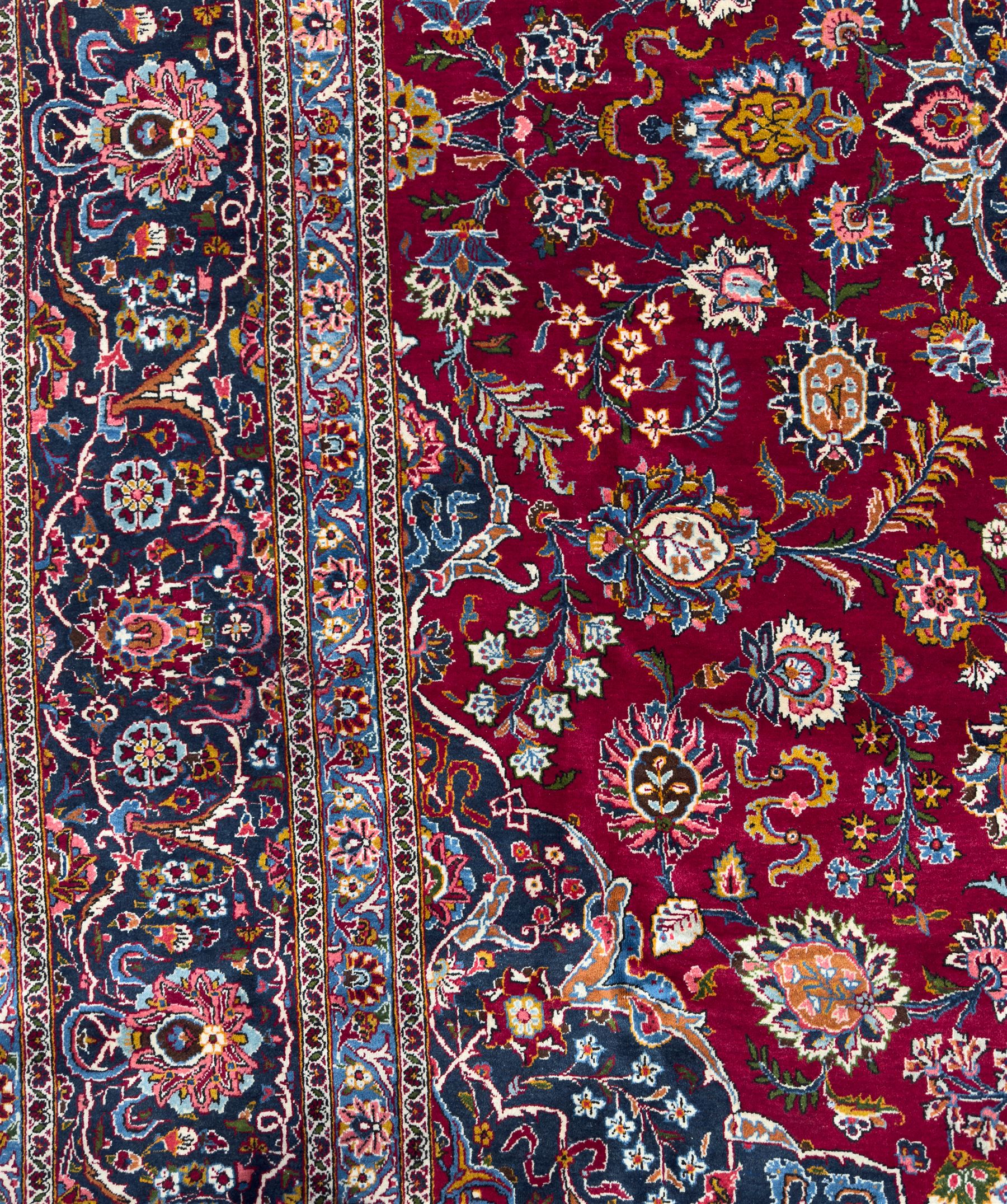 Central Persian Kashan hand-knotted carpet, madder red field with an intricate design of scrolling vines, palmettes, and rosettes with a central lobbed medallion with pendant motifs in shades of blue and ivory, and spandrels all within a blue-ground border of palmettes and flowerheads between multiple guard borders, 450cm x 335cm 