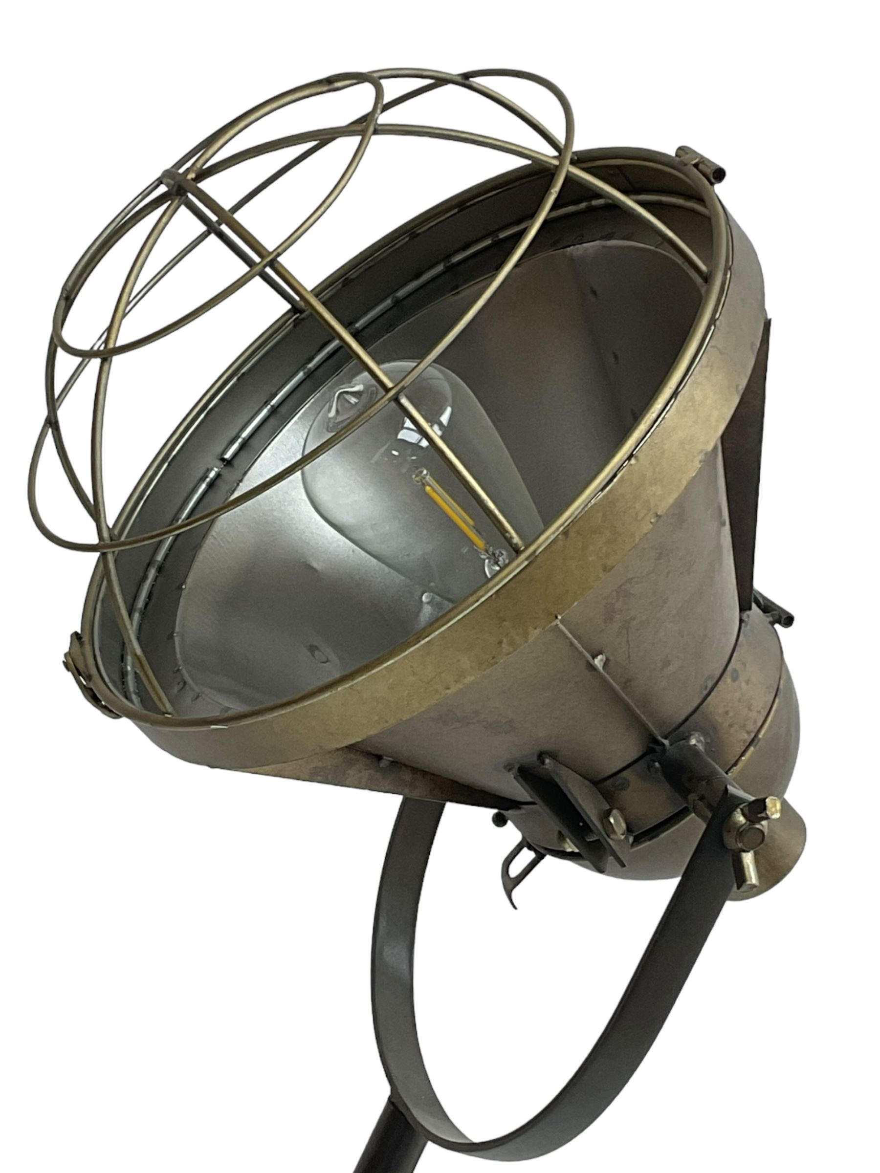 Industrial tripod spotlight, with a metal shade and wire guard, supported by a height-adjustable tripod base, the head equipped with an adjustable tilt mechanism