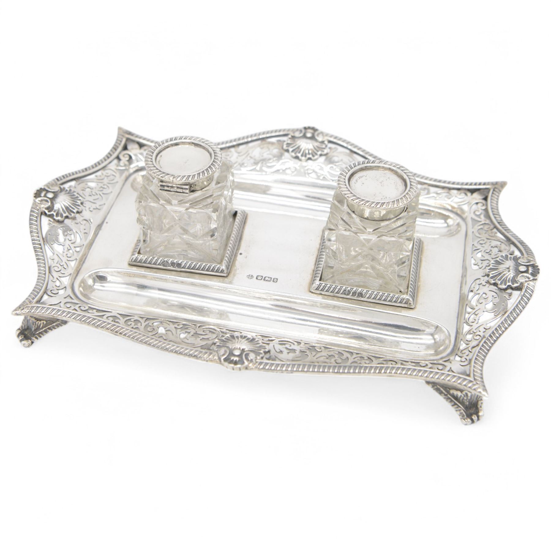Late Victorian silver desk stand, of rectangular form, with shaped and pierced, oblique gadrooned rim, upon four paw feet, the central tray with two pen holders and two reserves holding cut glass silver topped inkwells, hallmarked William Hutton & Sons Ltd, Sheffield 1900, tray W23cm