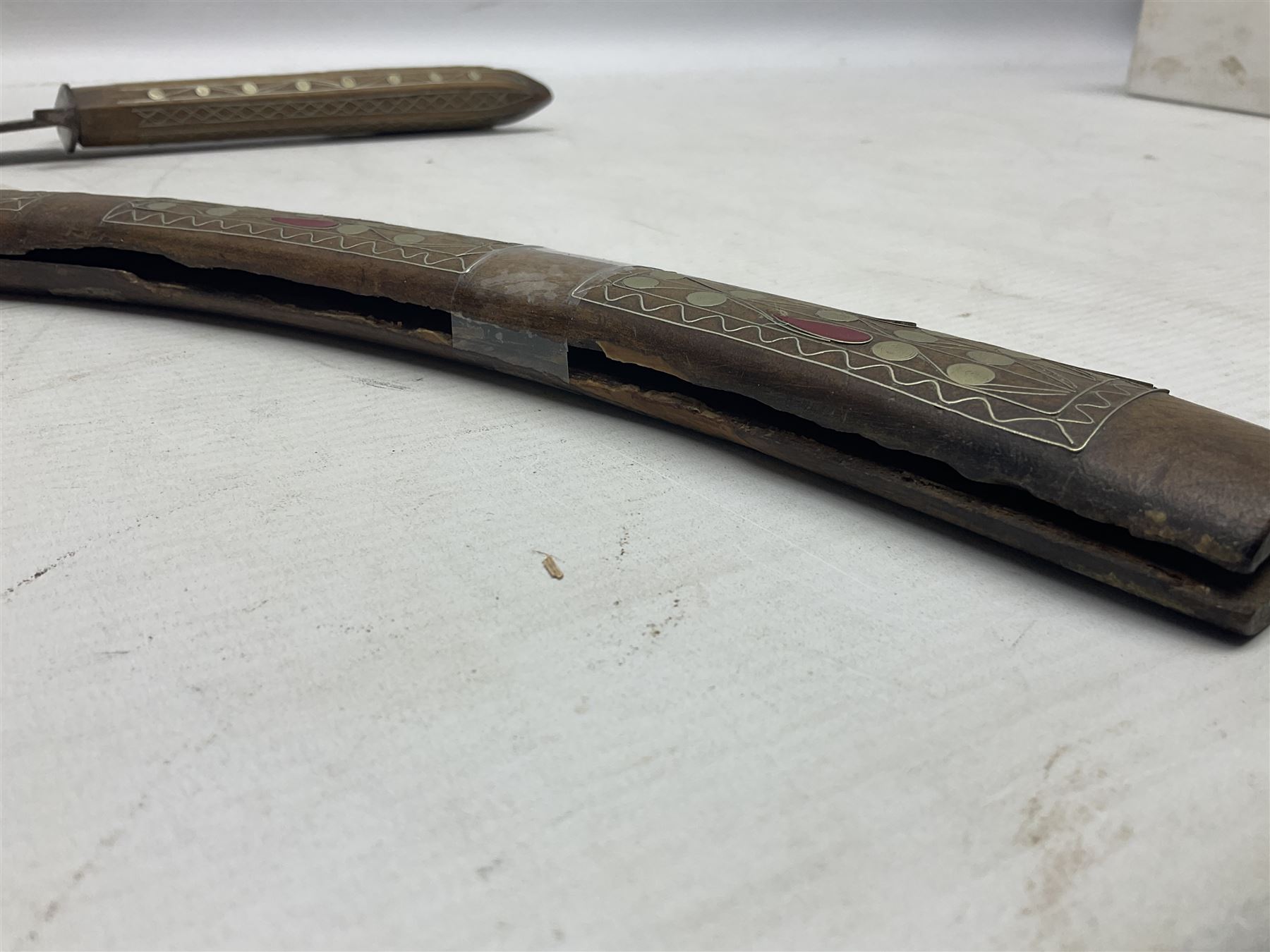 Late 19th/early 20th century Moroccan nimcha dagger-sword, the 40cm curving steel blade with engraved decoration on both sides; shaped hardwood hilt inlaid with brass, white metal wires and red coloured panels; in similarly inlaid scabbard L61cm overall