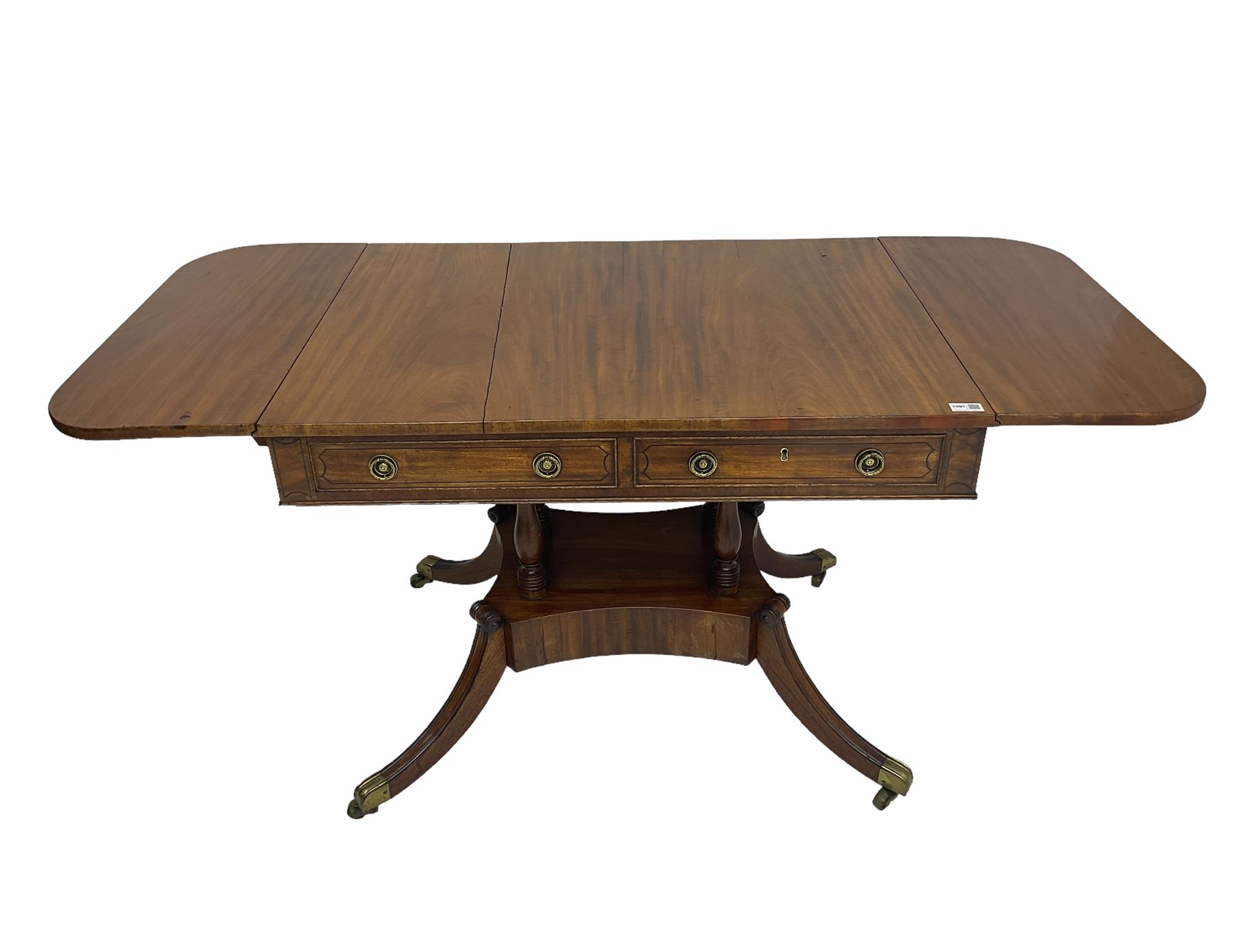 Regency period mahogany drop-leaf sofa table, rectangular top fitted with two cockbeaded drawers with ebony stringing, raised on turned columns united by quadrupod base, the sabre supports terminating in brass cups and castors