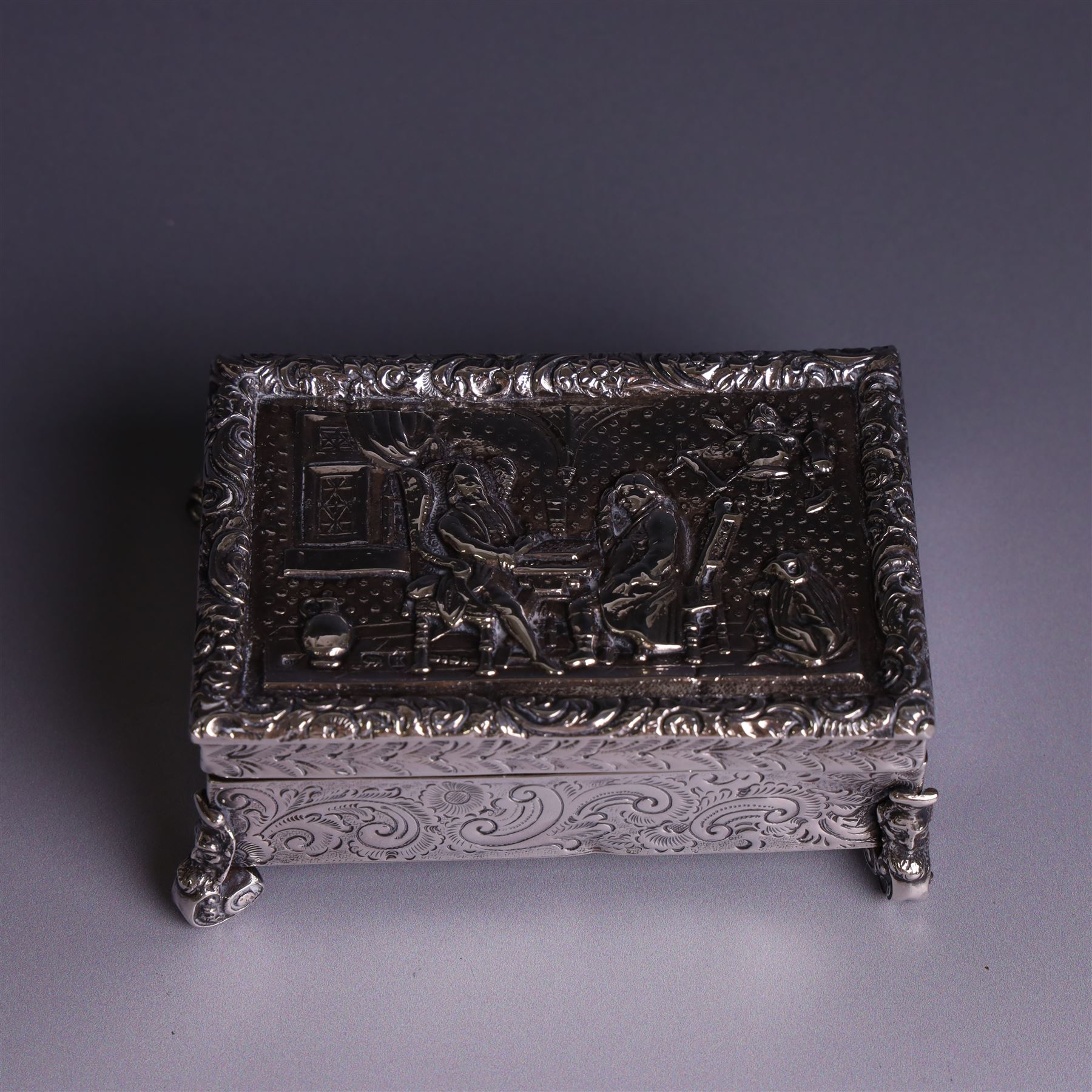 Victorian silver tobacco box, rectangular form, hinged lid embossed with a gaming scene, the body engraved with scrolls and flowers, upon four scrolling mask feet, hallmarked George Heath London 1885, H4.5cm, W10.5cm