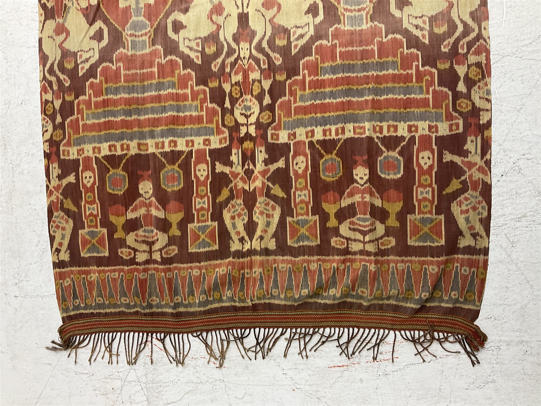 Ikat Man’s Cloth (Hinggi Kombu), East Sumba, Indonesia, woven in red, blue and yellow-brown warp ikat, constructed of two mirroring long panels, the motifs arranged in four bands depicting warrior figures astride rearing horses, high-ranking figures and guardians within geometric borders, on carved wooden wall hanger, L295cm
