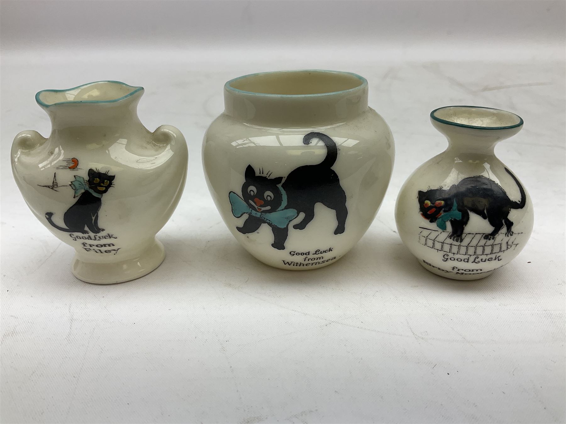 Five crested ware Good Luck black cats, comprising two Willow Art examples, two Arcadian examples and one Carlton China, to include Bridlington, Hull, Filey, etc, tallest H9cm
