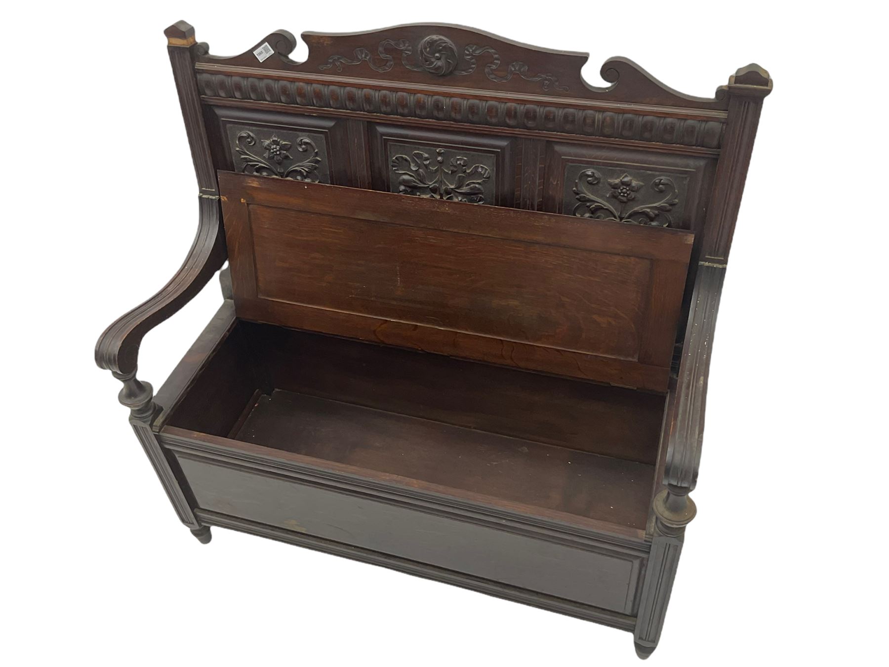 Late 19th century oak settle bench, shaped cresting rail carved with ribbon over triple panelled back, the panels relief carved with scrolled and floral urns and cartouche, hinged box seat, turned front feet, with upholstered squab cushion
