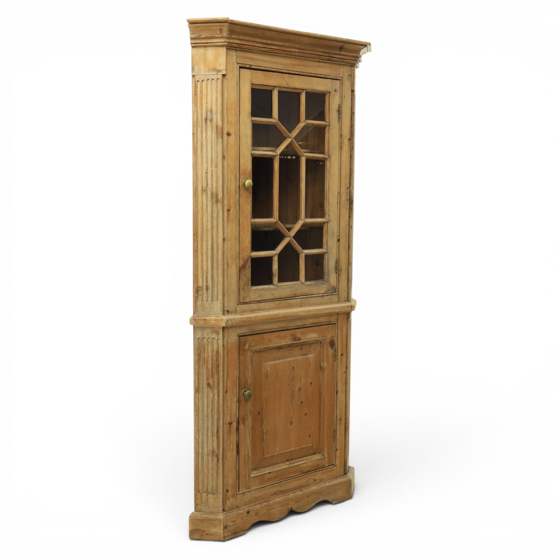 19th century stripped pine corner cabinet, projecting cornice flanked by fluted sides, astragal glazed upper door with brass knob, lower section with fielded panelled door, raised on a shaped plinth base 