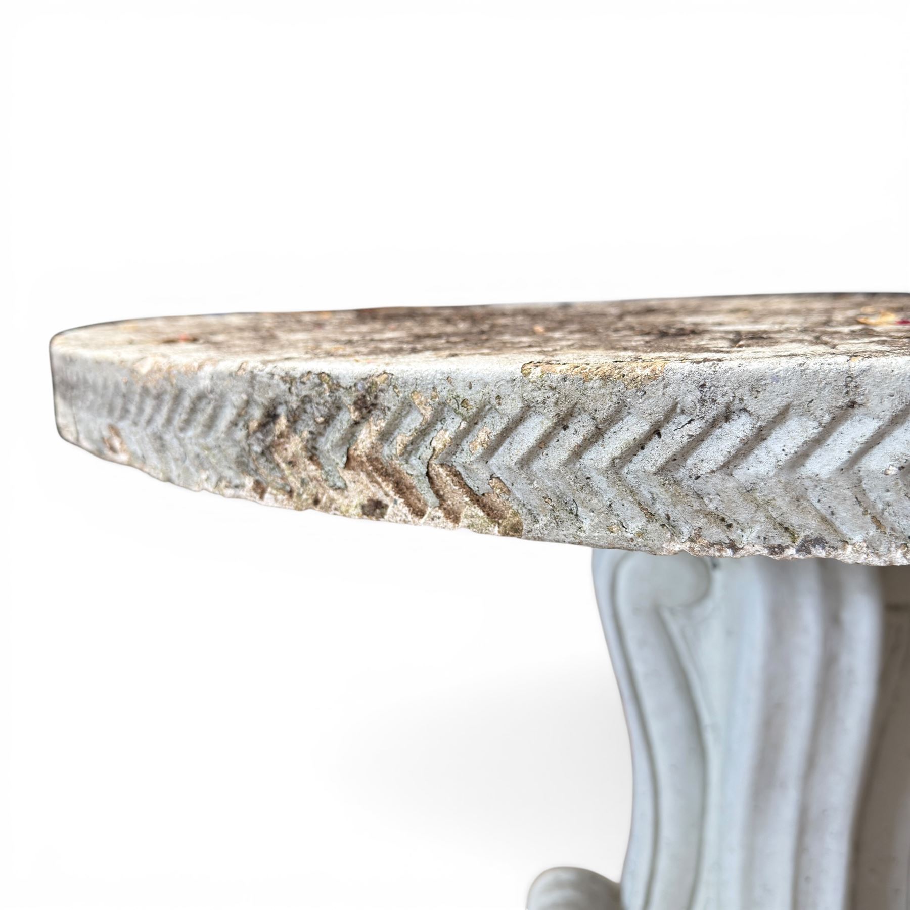 Weathered cast stone garden table, circular top etched with central floral medallion, the edge with repeating chevron motifs, raised on a quadriform S-scroll pedestal with stepped and chevron carved base