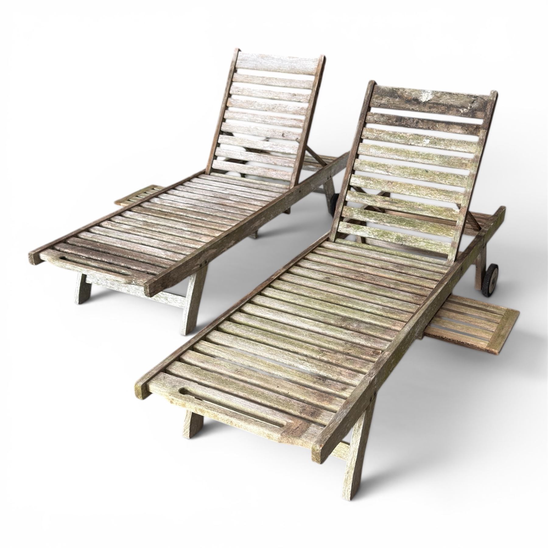 Pair of teak garden sun loungers, with slatted adjustable backrests, integrated wheels and sliding pull-out side trays, weathered natural finish.
