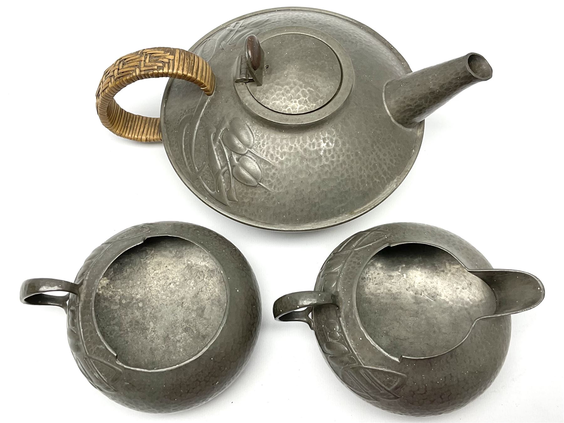 Archibald Knox for Liberty & Co Tudric pewter three piece tea service, comprising teapot with woven reed handle, single handled open sucrier, and milk jug, each with planished finish and detailed with honesty flowers, each impressed beneath Made in England Tudri Pewter 0231 