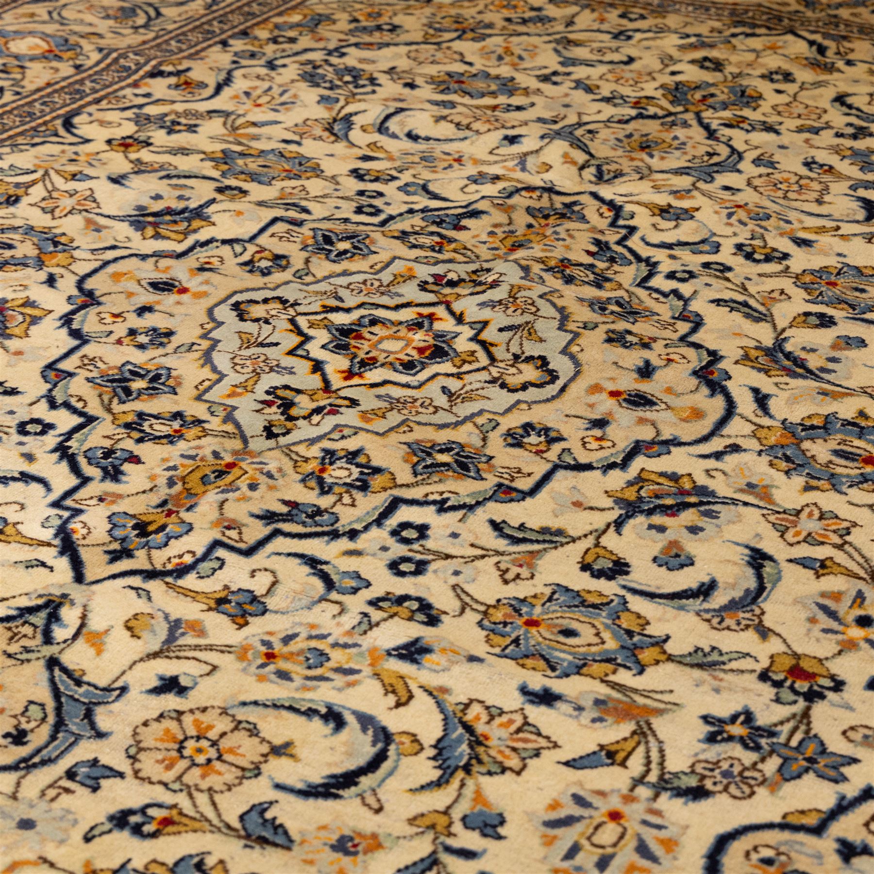 Central Persian Kashan hand-knotted carpet, ivory ground with central stepped floral medallion surrounded by scrolling vine work, palmettes, and stylized blossoms, field enclosed by a multi-border arrangement, wide main border decorated with repeating floral cartouches and foliate motifs, flanked by narrow guard stripes, with cotton fringe ends