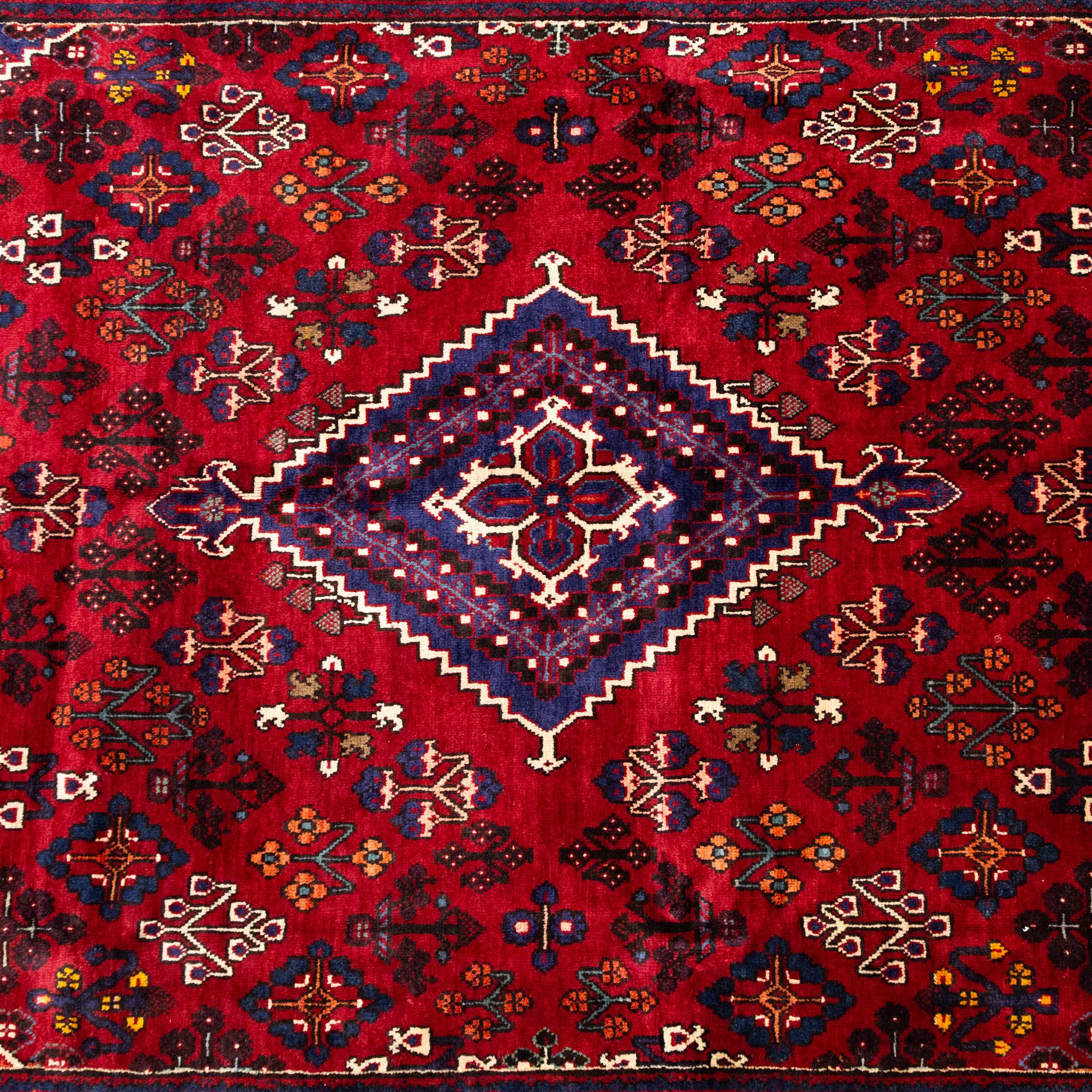 Central Persian Joshagan hand-knotted rug, madder-red field with central stepped lozenge medallion enclosed by angular spandrels, field decorated with repeating stylized geometric floral motifs and small diamond devices enclosed by a dark border of geometric ornament between multiple guard stripes, with cotton fringe ends  