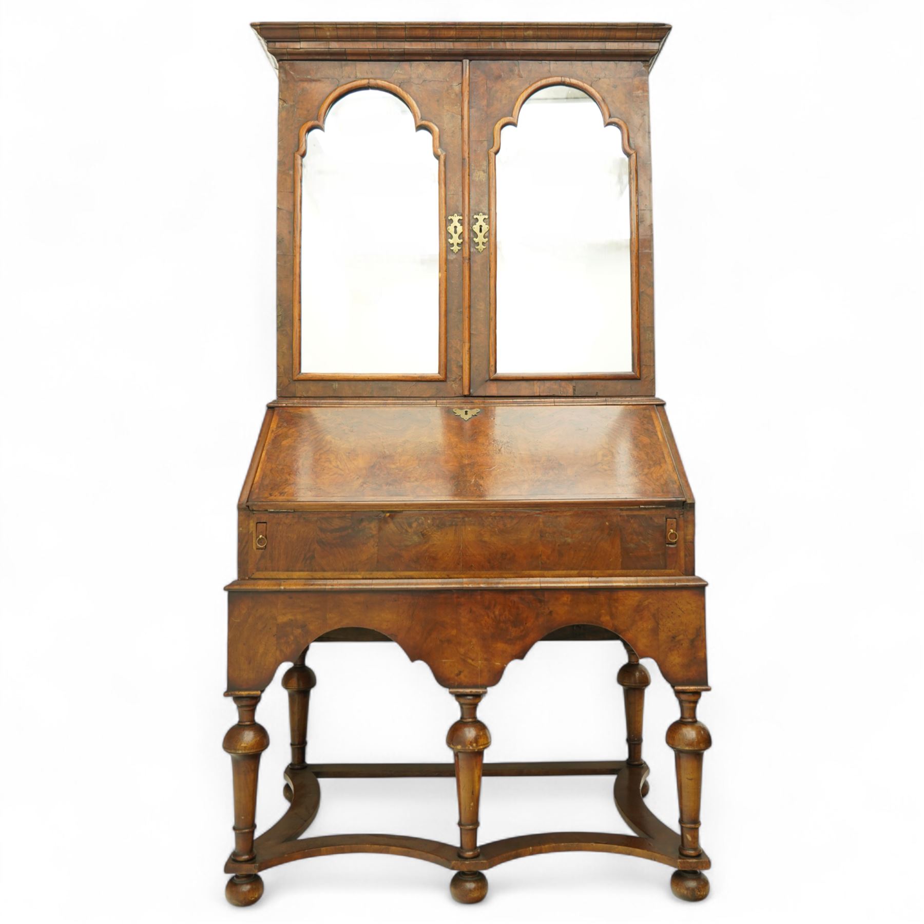 Queen Anne burr walnut chest on stand, moulded projecting cornice over arched mirror glazed doors, above a sloping fall front enclosing fitted interior with pigeonholes and correspondence drawers, fitted with foliate brass escutcheons, shaped apron over tapered baluster turned supports united by curved stretchers, early 18th century with later restorations 