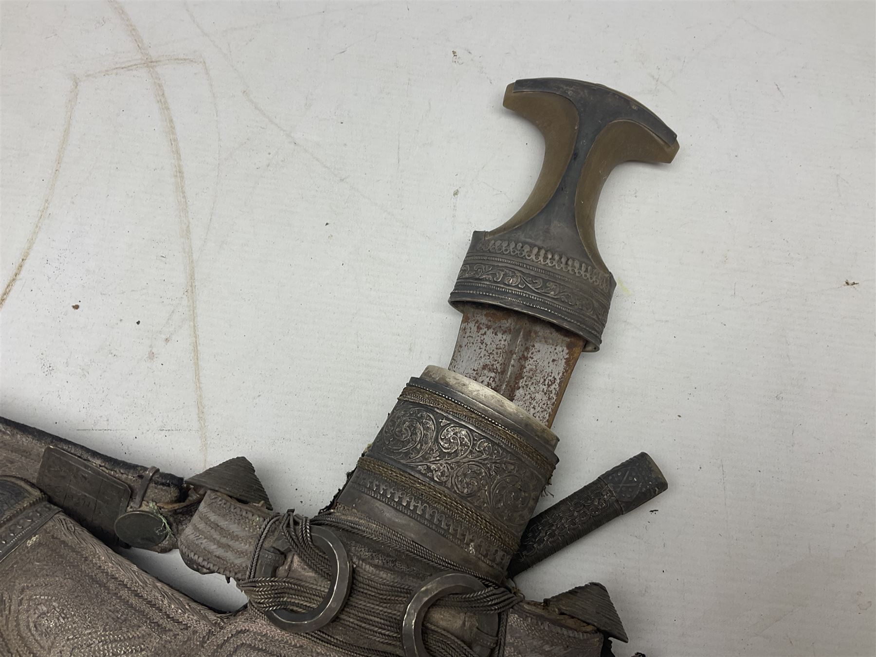 Omani Khanjar dagger, the 19cm curved blade with pronounced medial rib to either side, waisted hilt with dome shaped pommel and silver mounted horn grip decorated with an embossed band; in an intricately decorated silver scabbard with silver rings woven into the belt mount and fitted with original coin mounted silver thread-work belt with additional side knife in sheath verso L34cm overall, belt length 81cm; together with a Nepalese kukri in leather scabbard with one skinning knife (2)