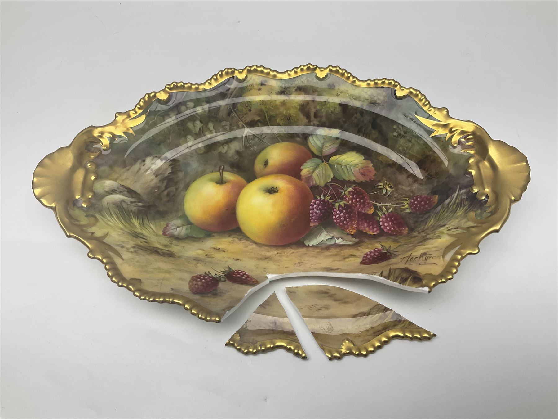 Early 20th century Royal Worcester dish decorated by Thomas Lockyer, oval form with gilt shaped rim and twin shell handles, hand painted with a still life of fruit upon a mossy ground, signed T Lockyer, with puce printed mark beneath and date code for 1922, for restoration, W31cm