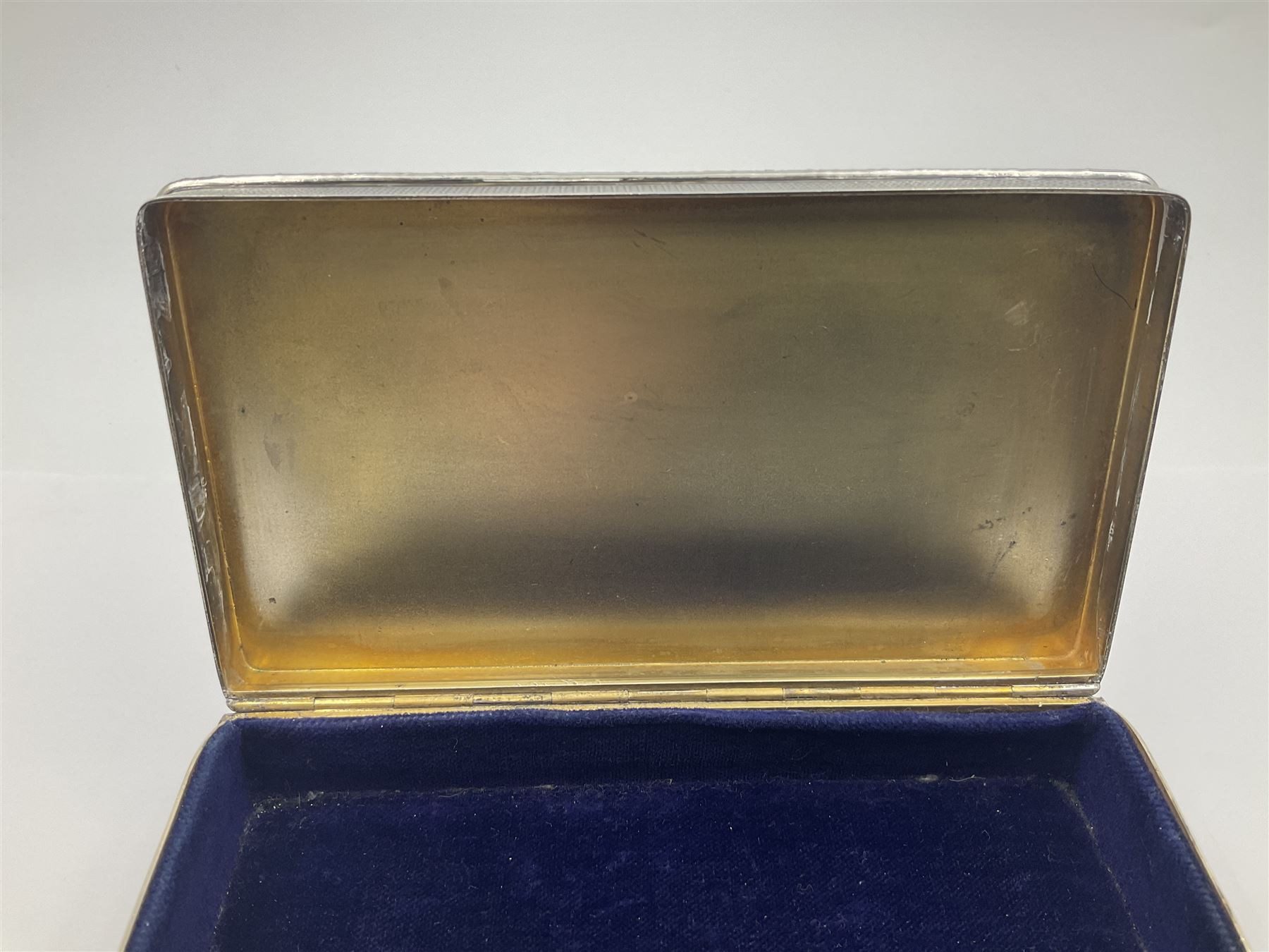 1920's silver table cigarette box, of rectangular form with engine turned decoration to the exterior, the hinged cover with central vacant rectangular panel and raised foliate rim, opening to reveal a gilt interior with later plush lining, hallmarked Charles Henry Dumenil, London 1923, H3.5cm L14.5cm D8.5cm